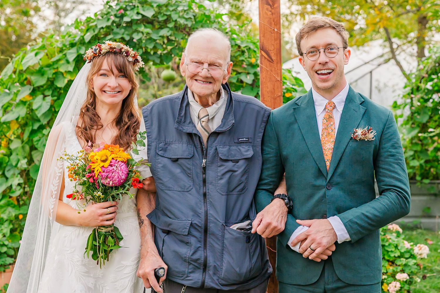 A wedding couple and an older relative pose together in a garden setting with bright autumn flowers.
