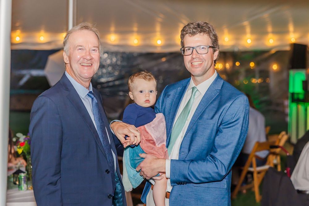 Three generations pose together at an indoor event with warm lighting and decorative elements in the background.
