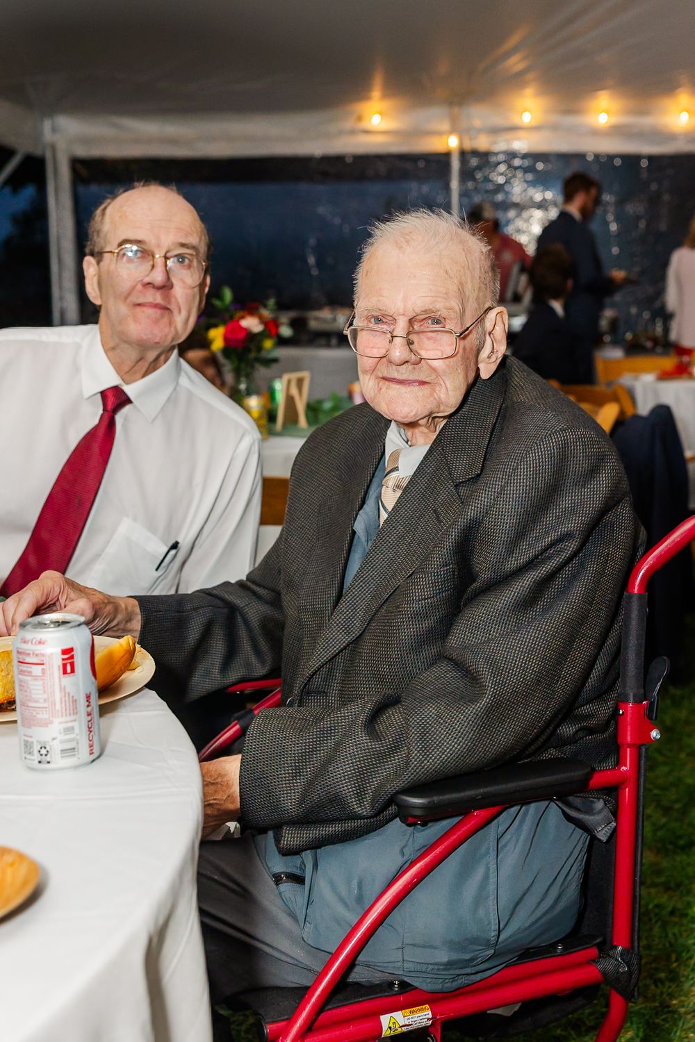 Elderly man in gray suit and glasses sits in red chair at outdoor event with refreshments on table.