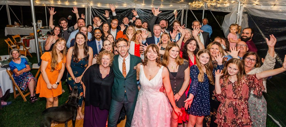 Large group photo of wedding guests celebrating together under a tent with festive lighting.