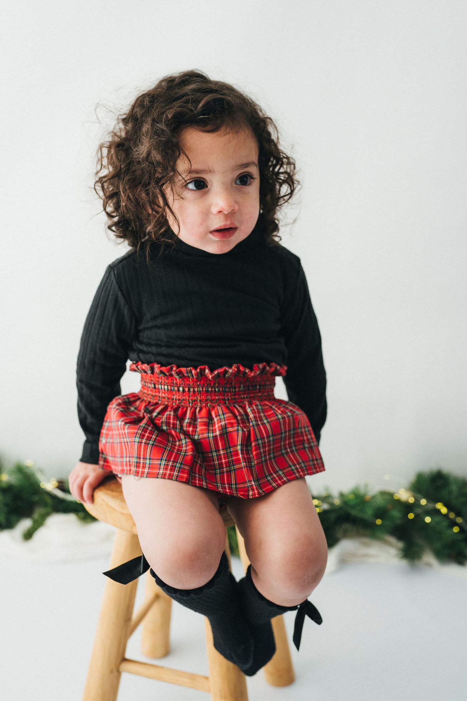 Petite fille assise sur un tabouret, vêtue d’une jupe écossaise rouge et d’un pull noir, lors d’une mini-séance photo Noël au studio Mon