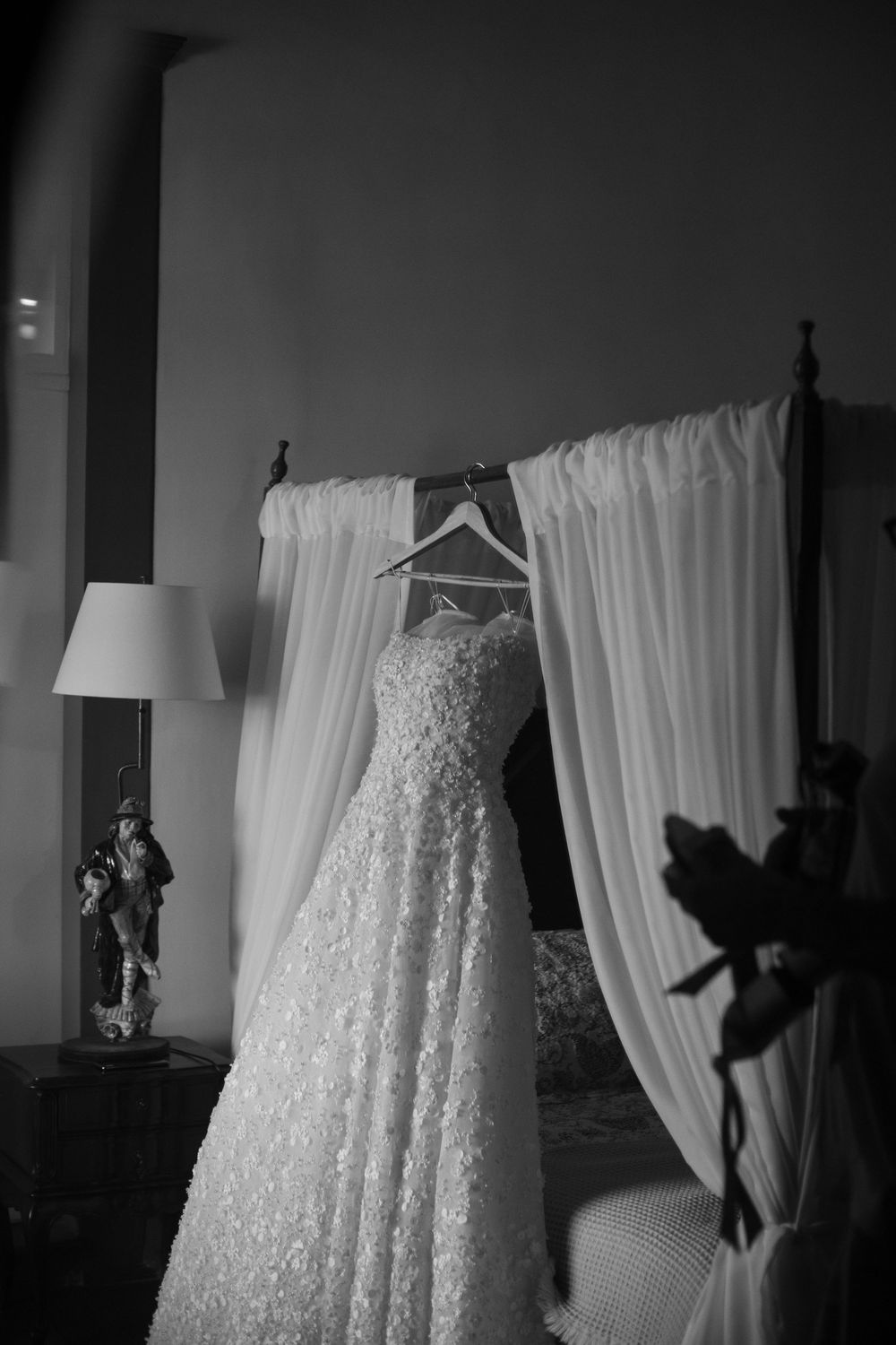 A white lace wedding dress hanging in front of sheer curtains in dramatic lighting.