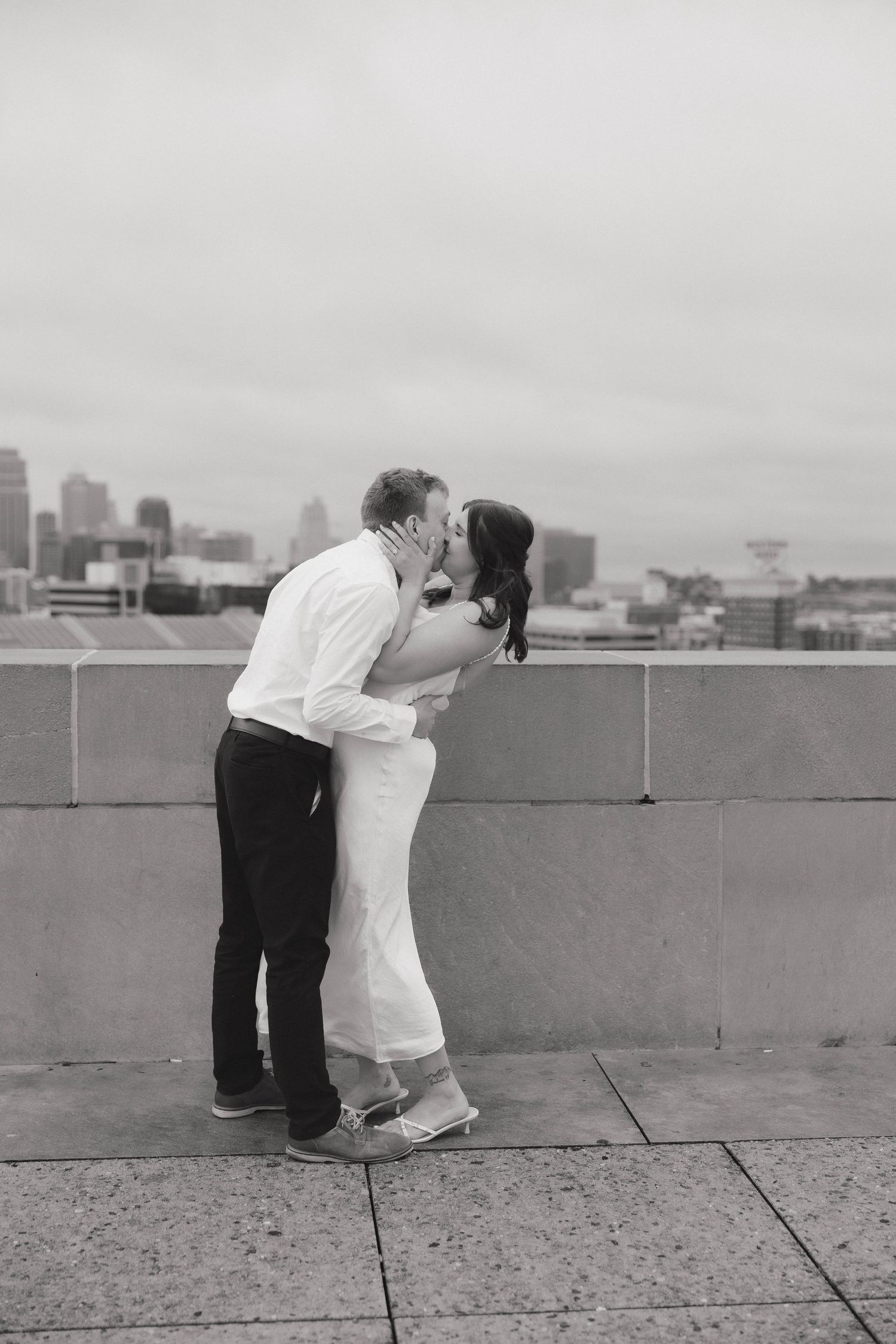 A couple shares a romantic kiss on a rooftop with a city skyline in the background in black and white.