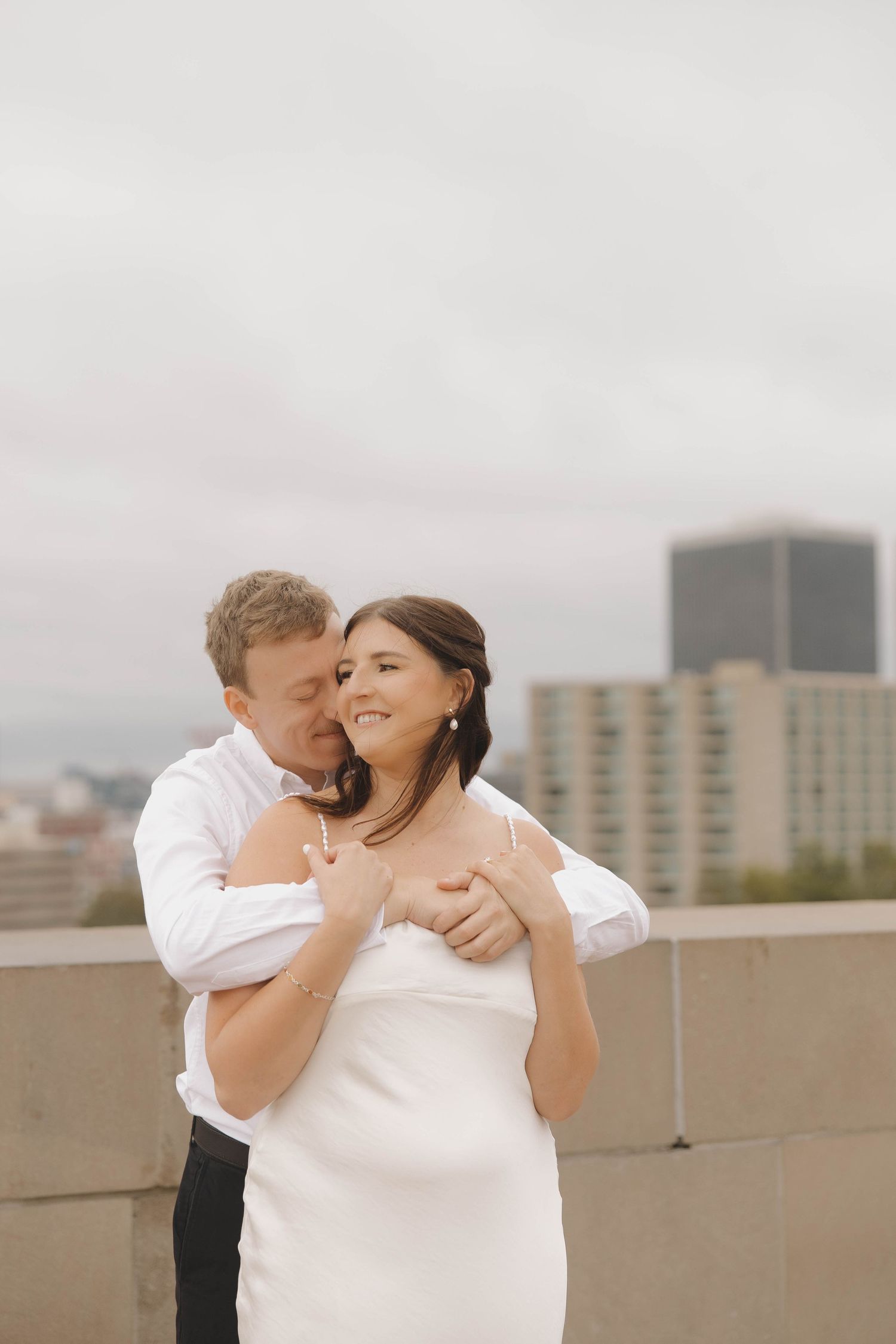 A romantic embrace between a couple wearing white and black formal attire on a rooftop with city buildings in the background.