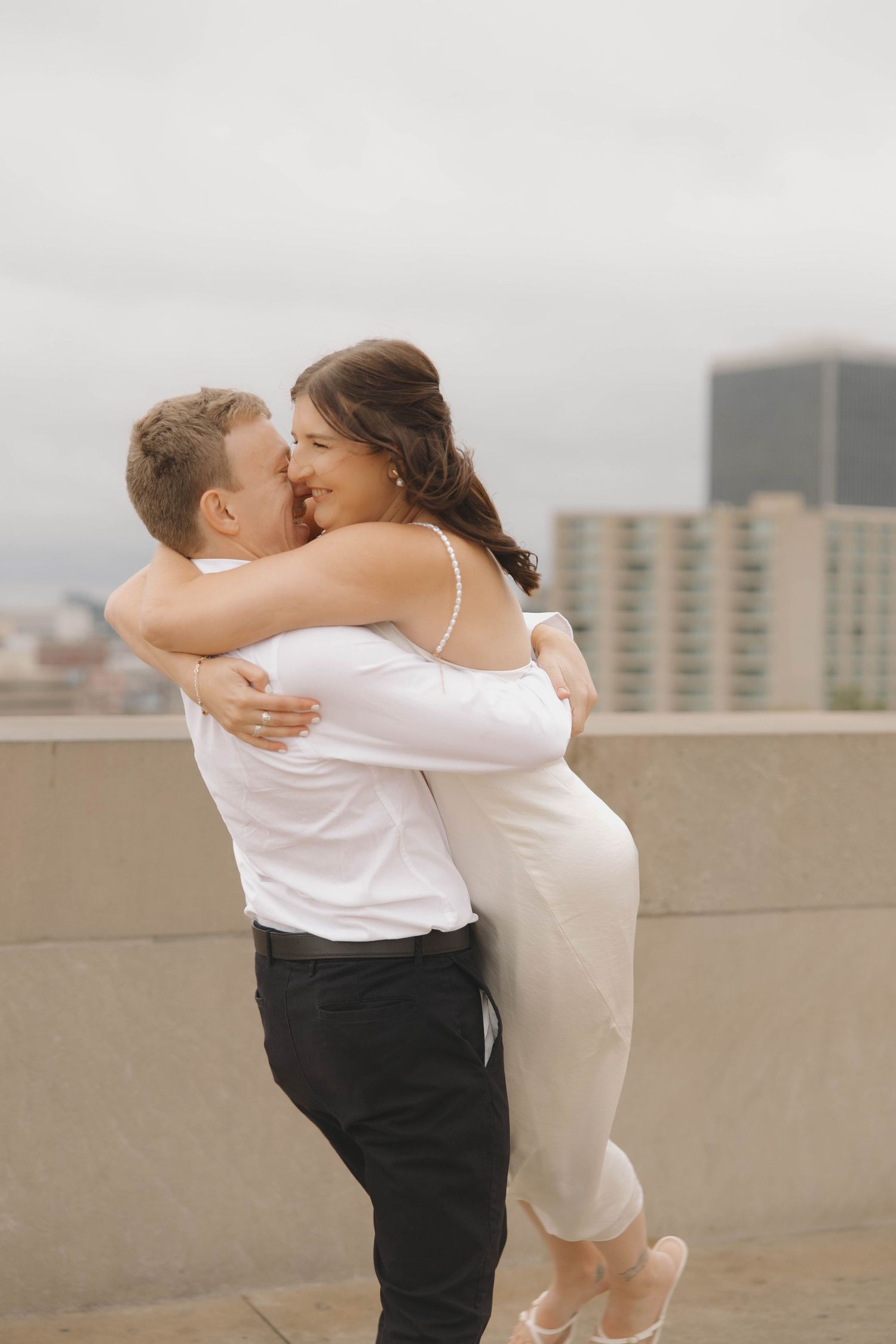 A romantic couple in formal attire share an intimate moment on a rooftop with city buildings in the background.