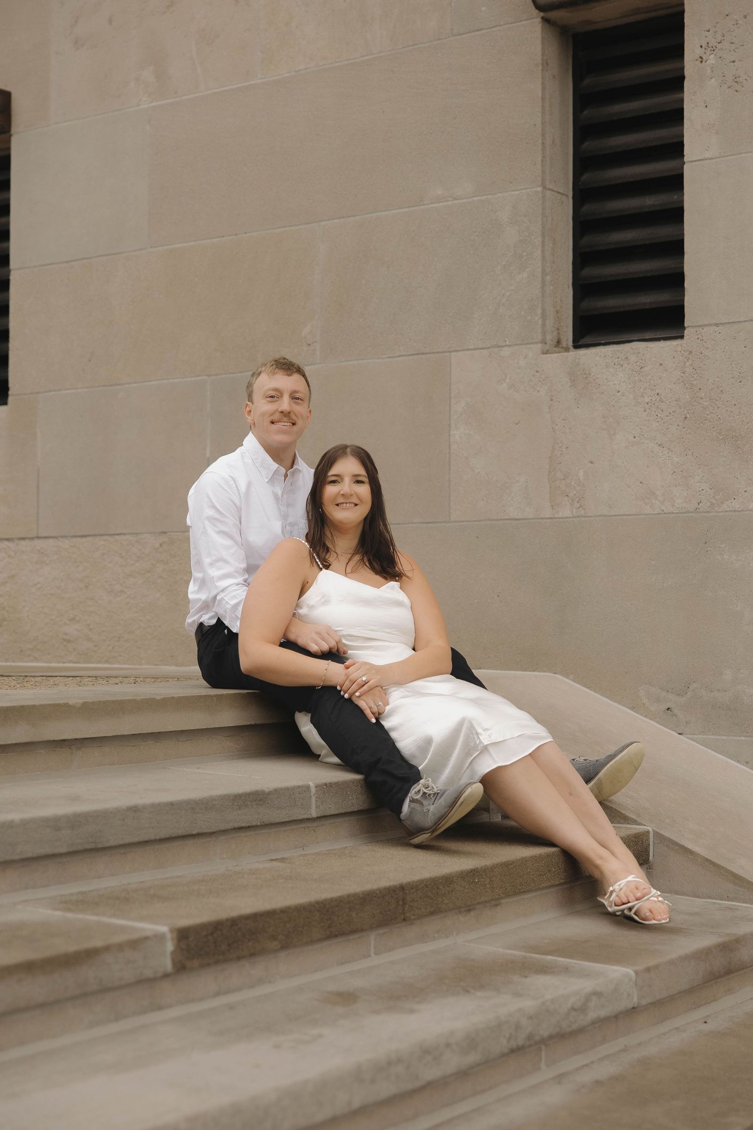 A couple in wedding attire sitting and embracing on concrete steps of a modern building.