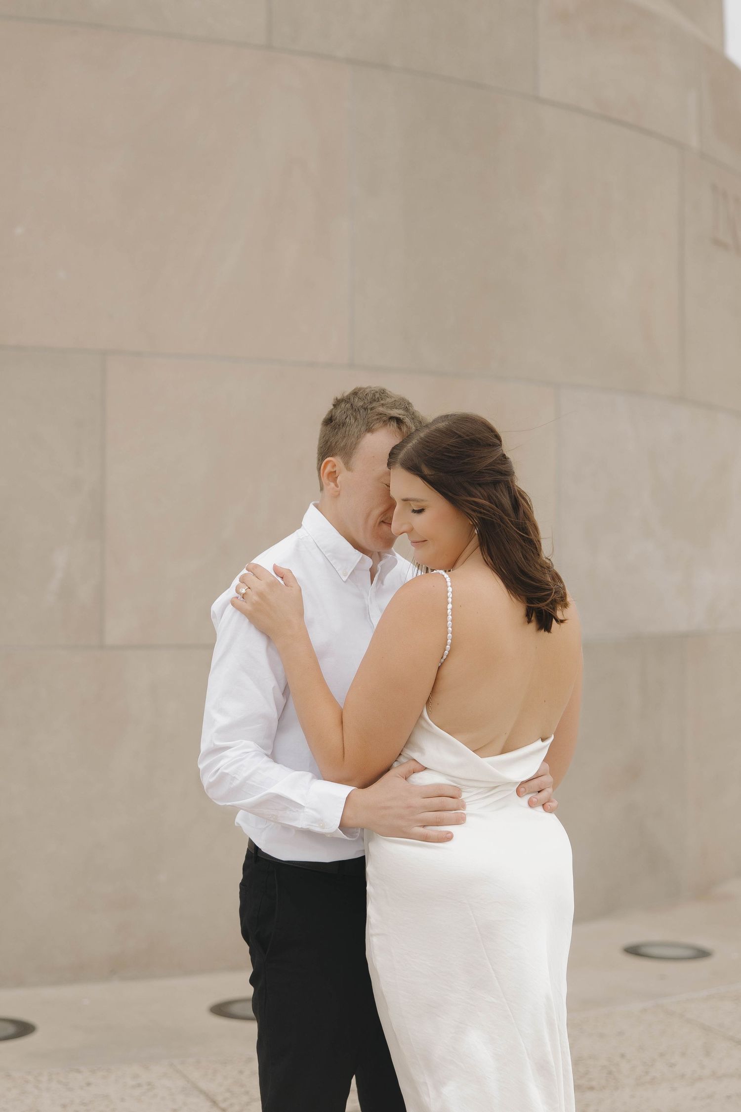 A couple in wedding attire embrace tenderly against a minimalist concrete wall backdrop.