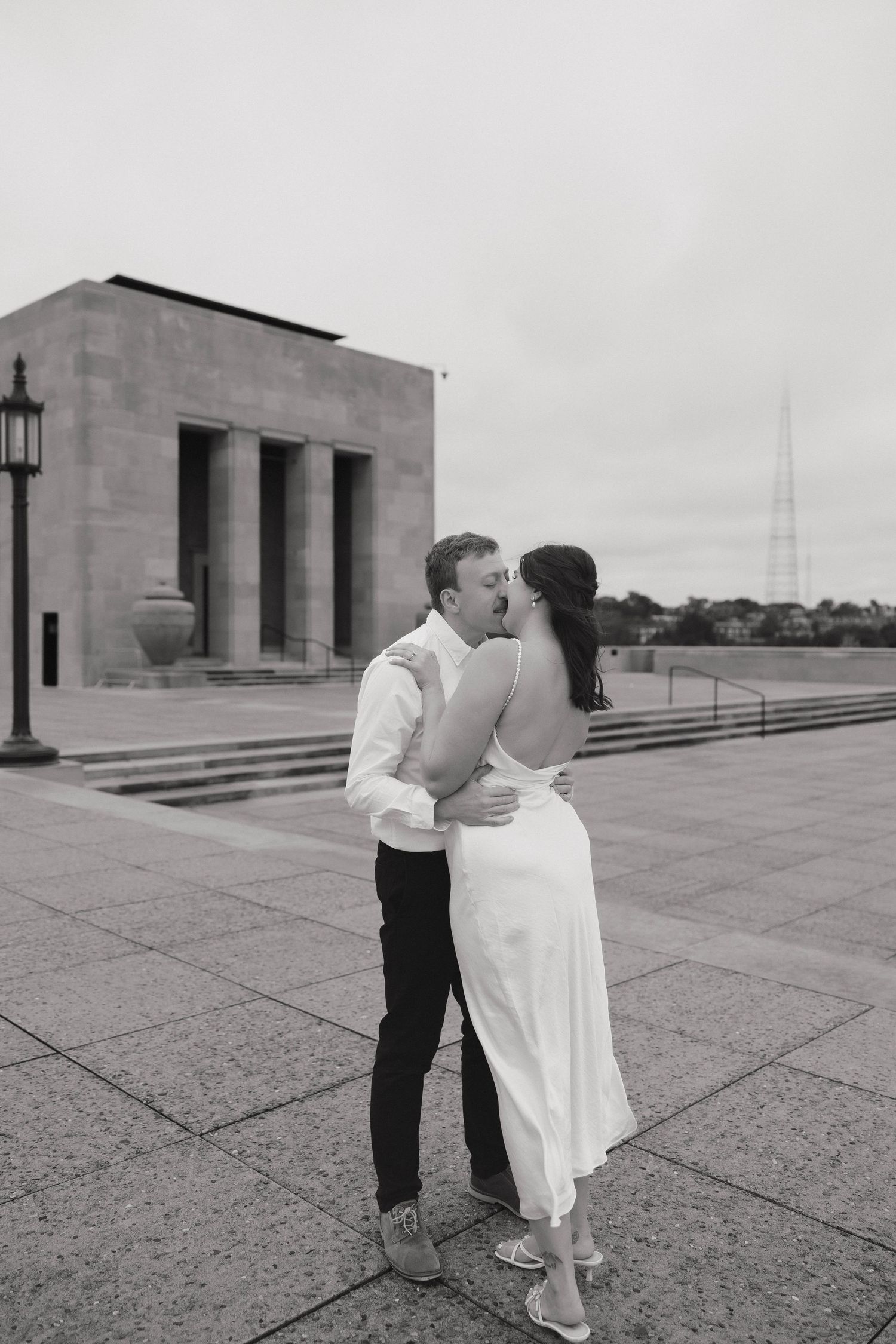 A black and white photo captures a couple dancing on a concrete plaza with modern architecture.