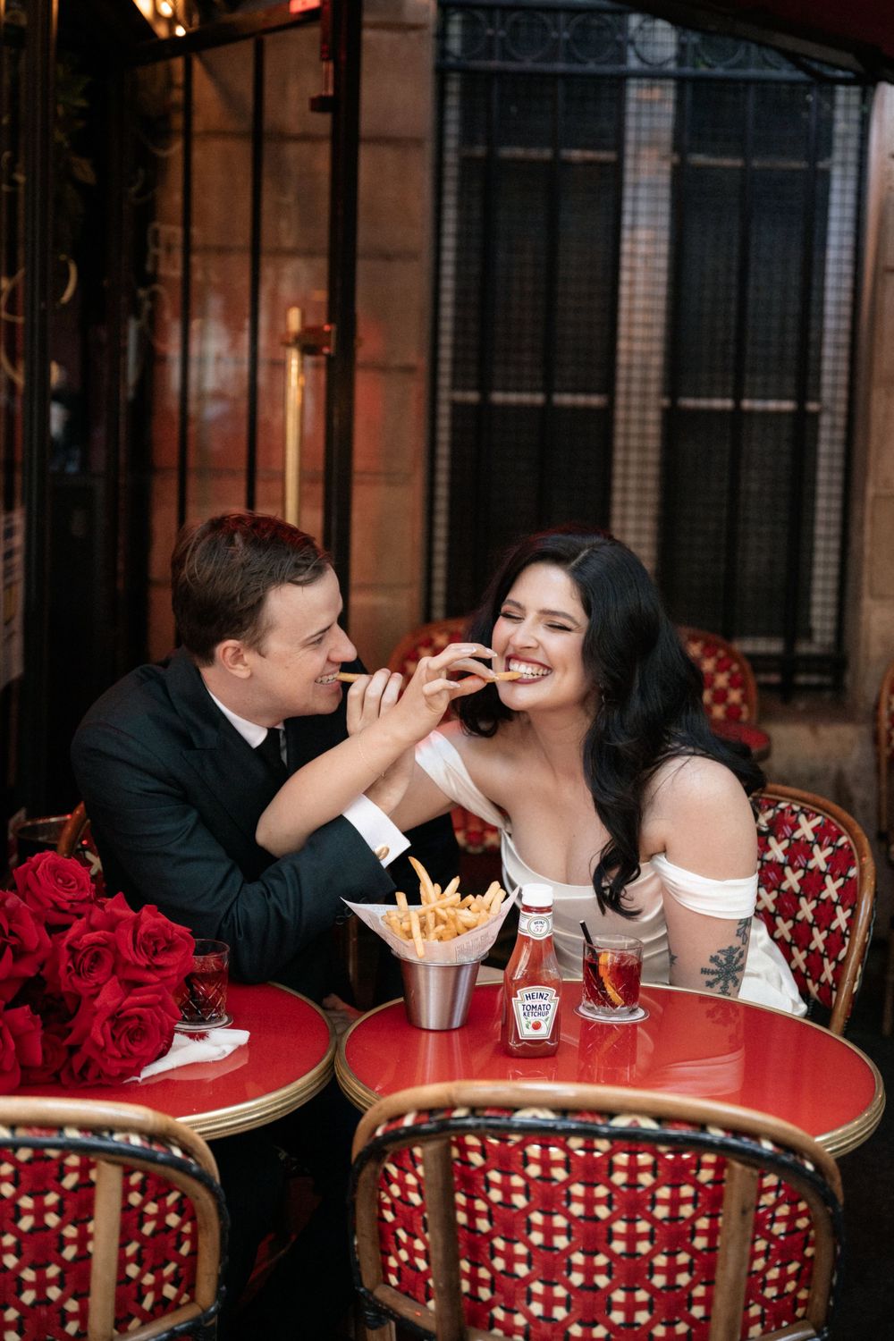 A romantic couple shares french fries at an outdoor Parisian cafe with red chairs and floral decor.