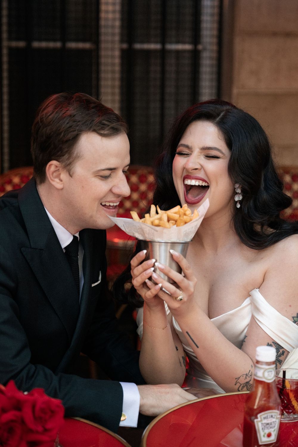 A couple sharing french fries and laughing together during their wedding reception.