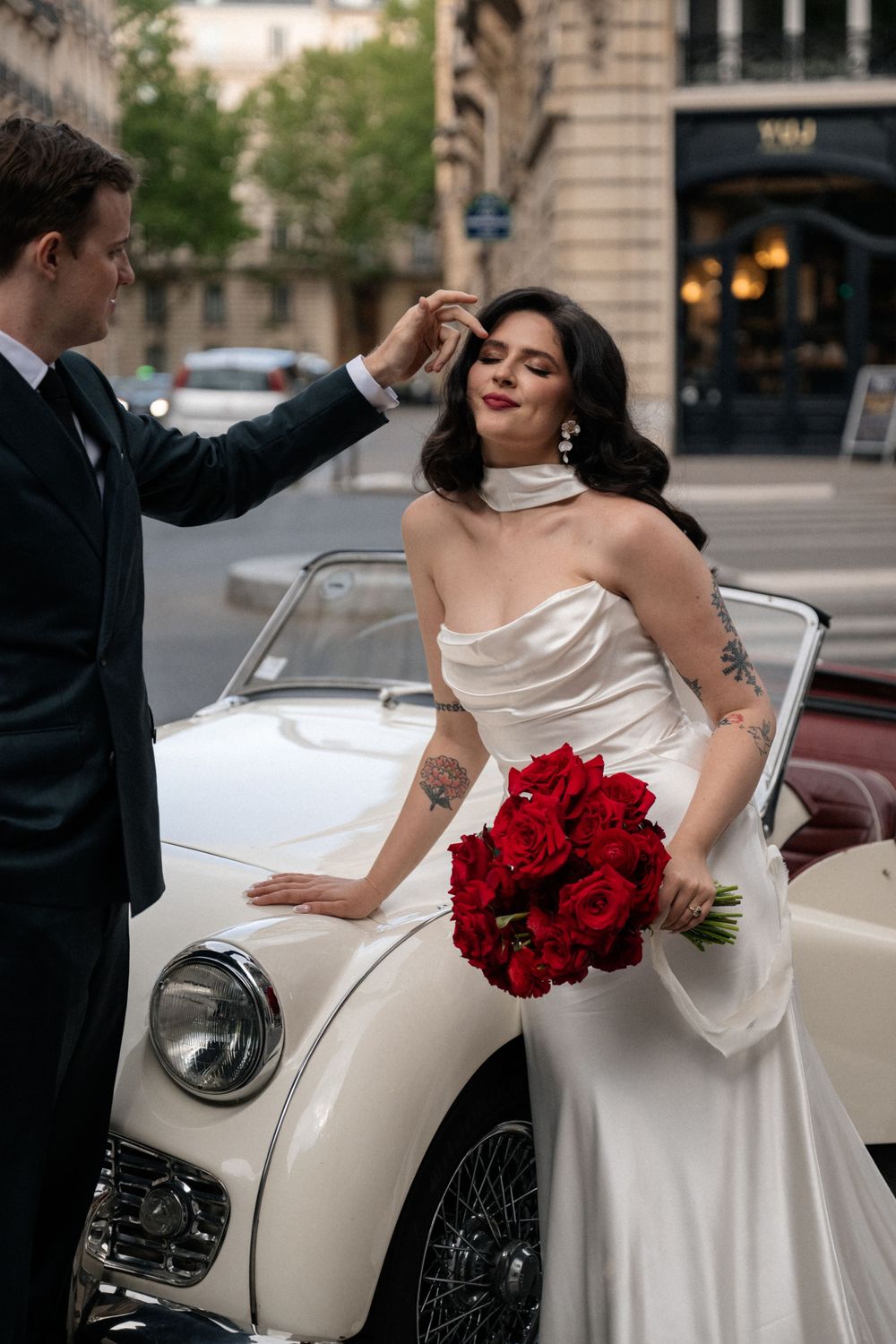 A couple poses with a vintage white car while holding a red bouquet in an elegant setting.