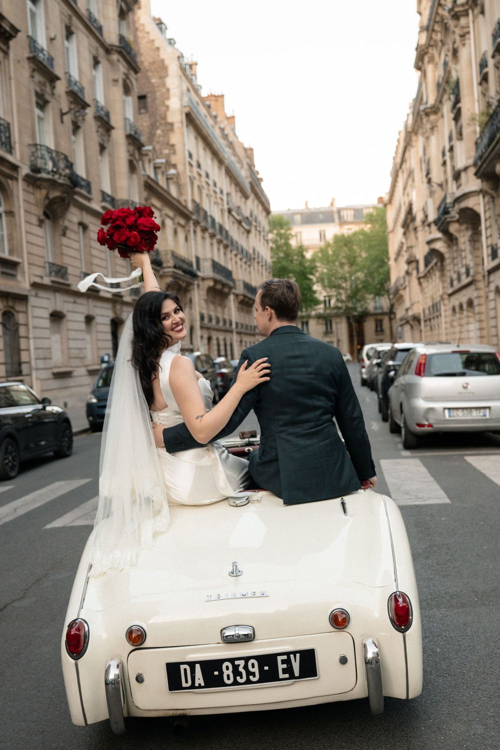 Newlyweds riding in a vintage white convertible car through Parisian streets with red roses.