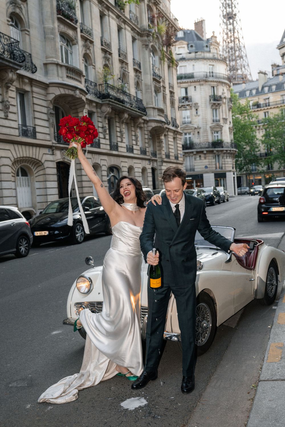 A celebratory moment captured on a Parisian street with a vintage car and red bouquet.