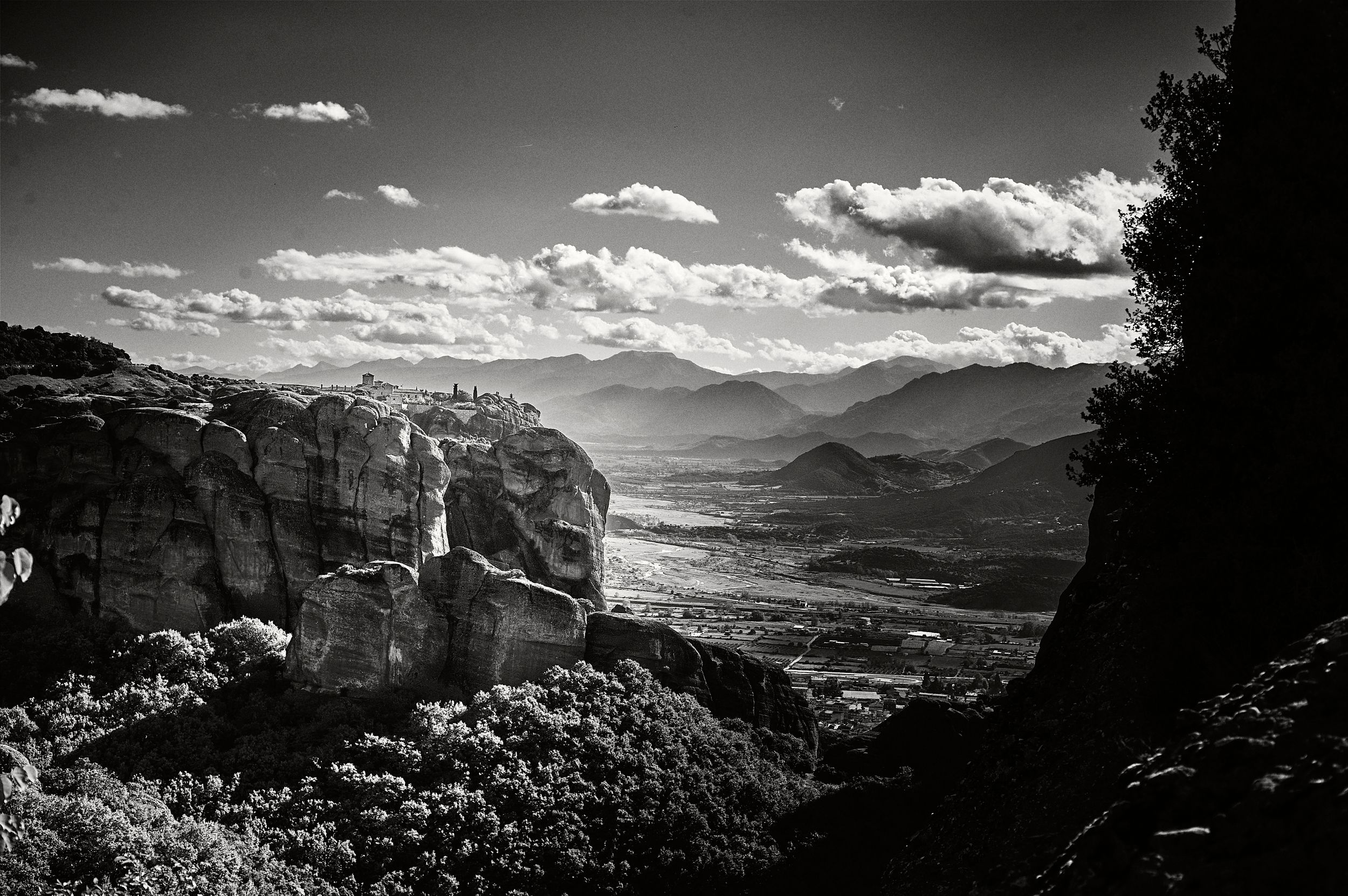 A sweeping black and white landscape of Meteora's rocky cliffs and monasteries overlooking a vast valley below.