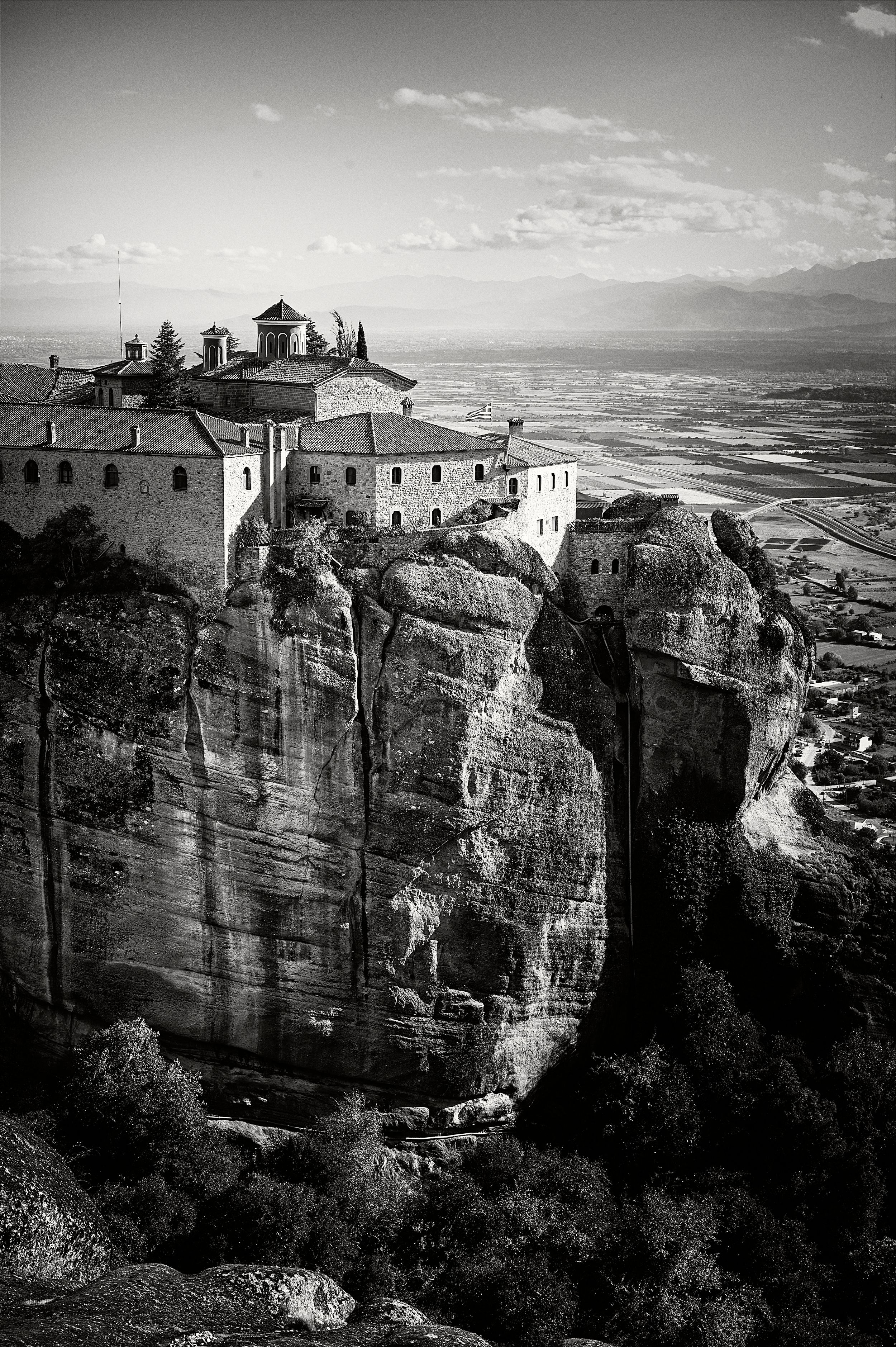 Dramatic black and white view of a monastery perched atop steep cliffs at Meteora, Greece against a cloudy sky.