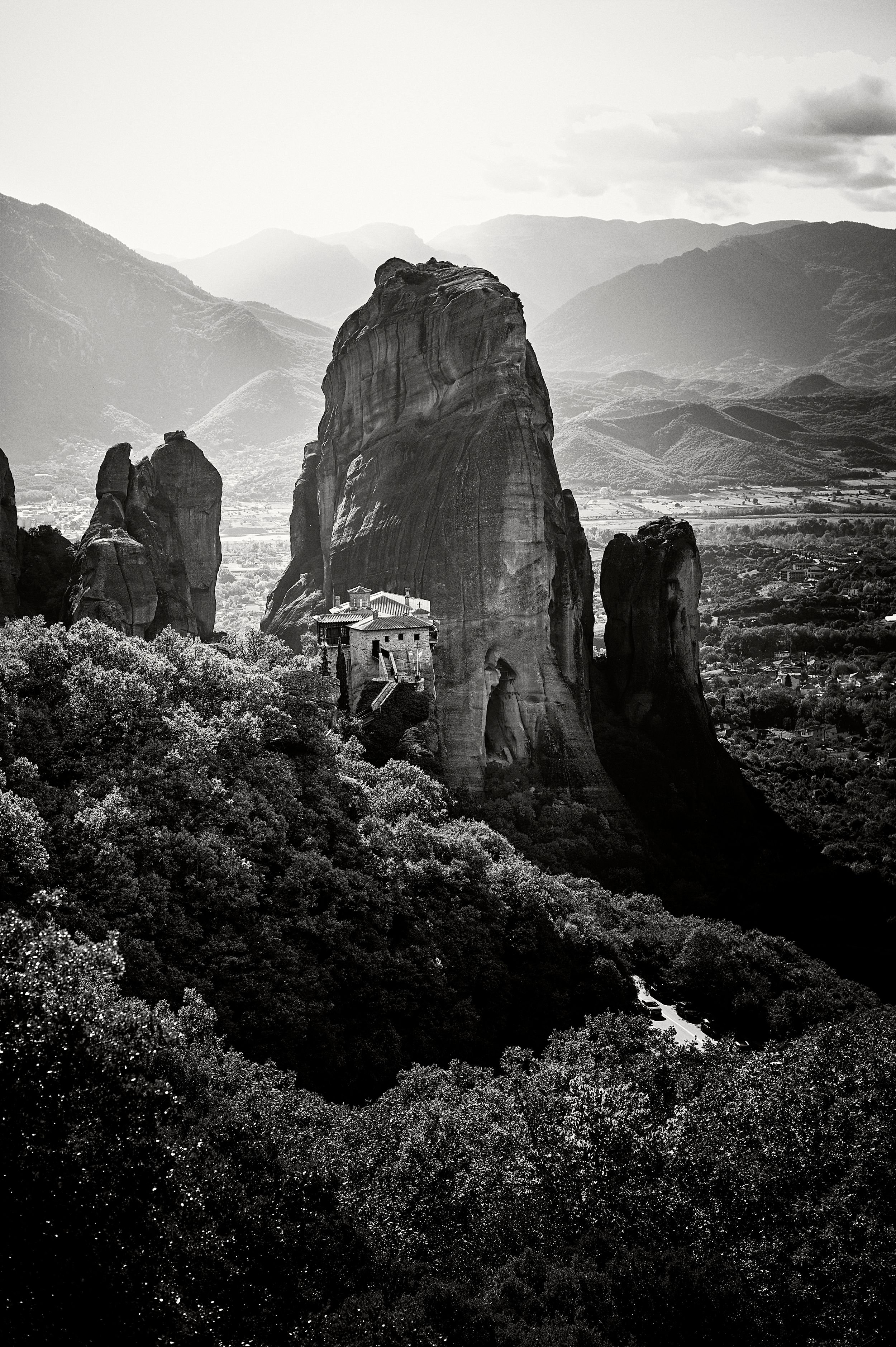 Dramatic black and white landscape of towering rock formations at Meteora, Greece with mountains in the background.
