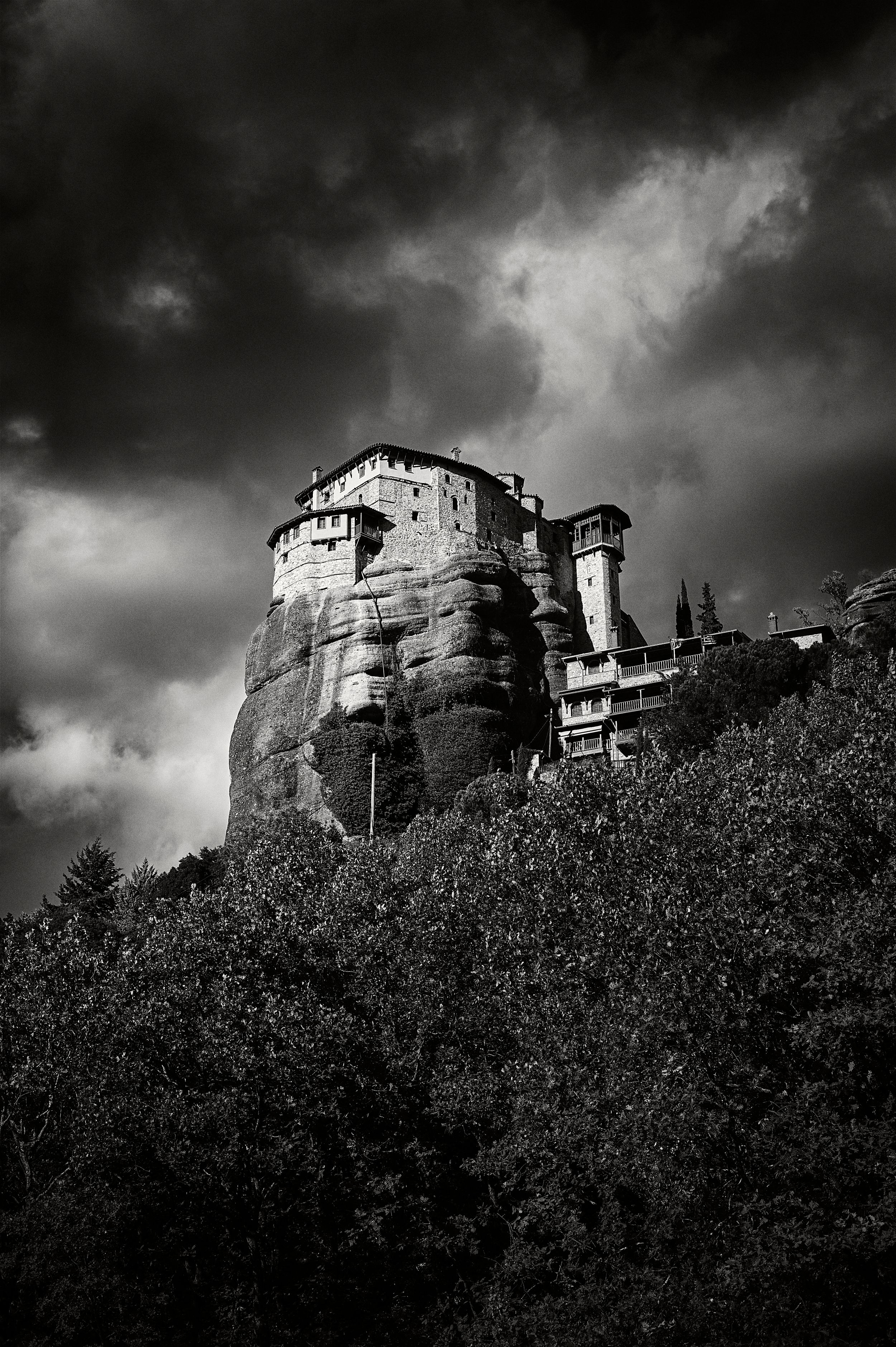 Dramatic black and white panorama of ancient Meteora monasteries perched on rocky cliffs under stormy skies in Greece.