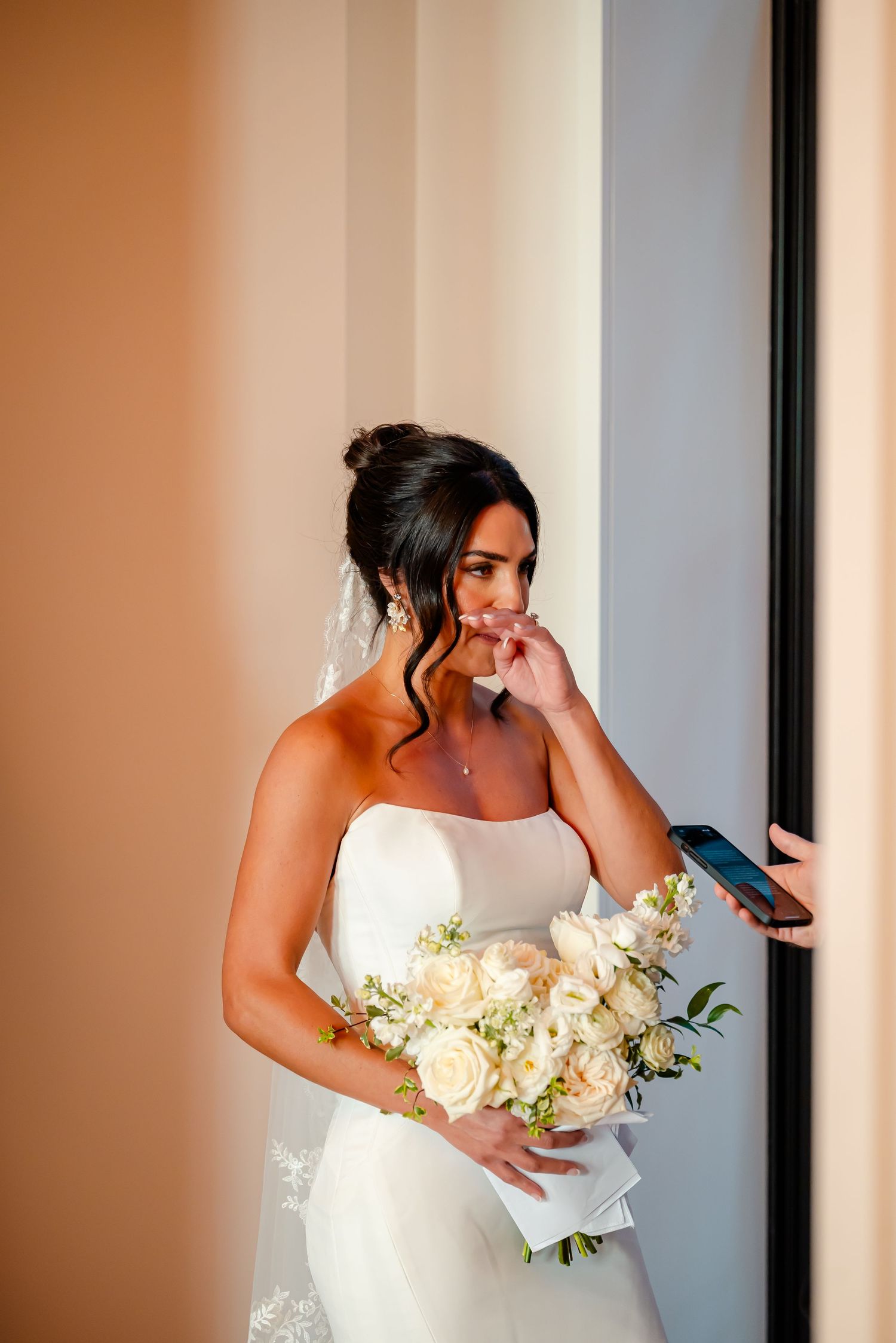 A bride in a white wedding dress holds a bouquet of white flowers while showing emotion during a first look moment.
