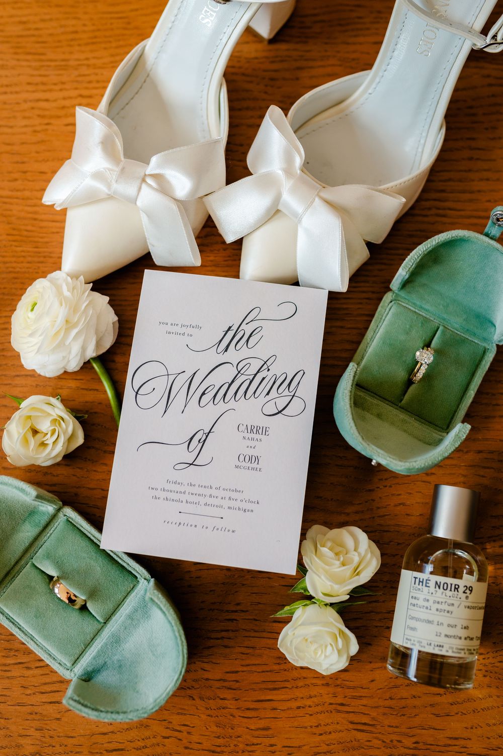 Wedding invitation with white bows, green velvet shoes, and cream roses arranged on wooden surface.