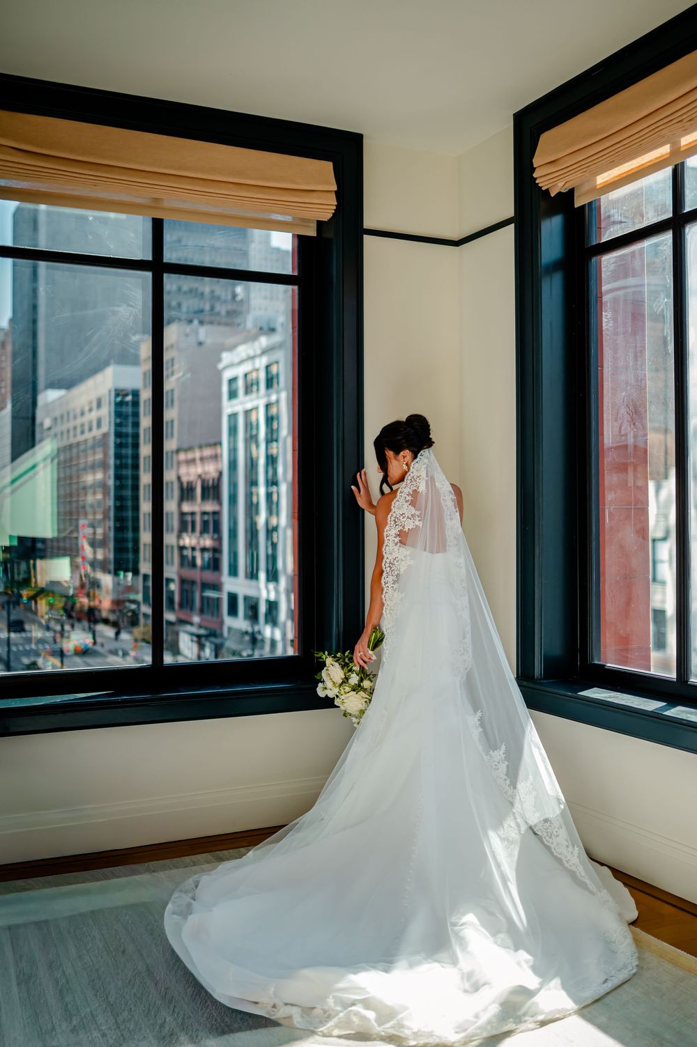 A flowing white wedding dress with a long cathedral veil is photographed against large windows in a bright room.