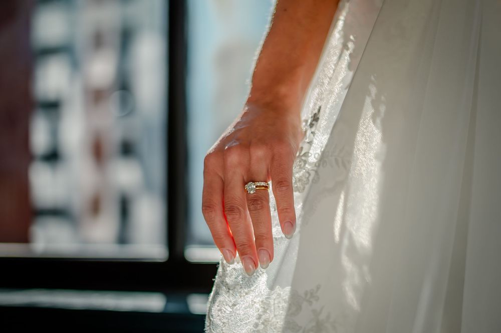 Close-up of a bride's hand wearing an engagement ring against white lace wedding dress in natural light.