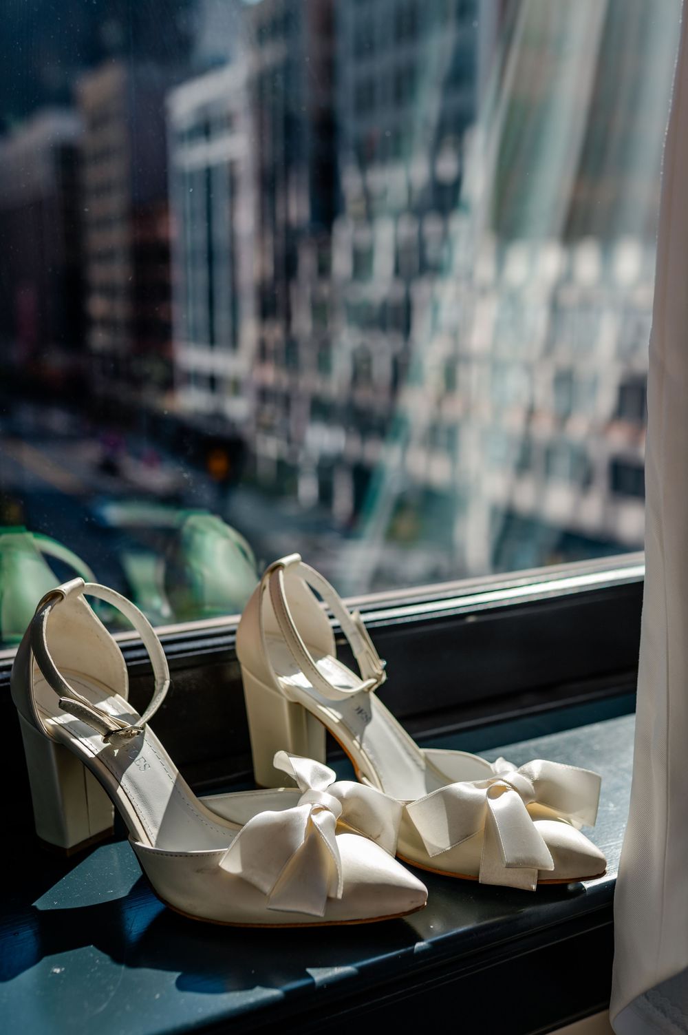 White bridal shoes with bows sit elegantly on a window ledge against a blurred city backdrop.