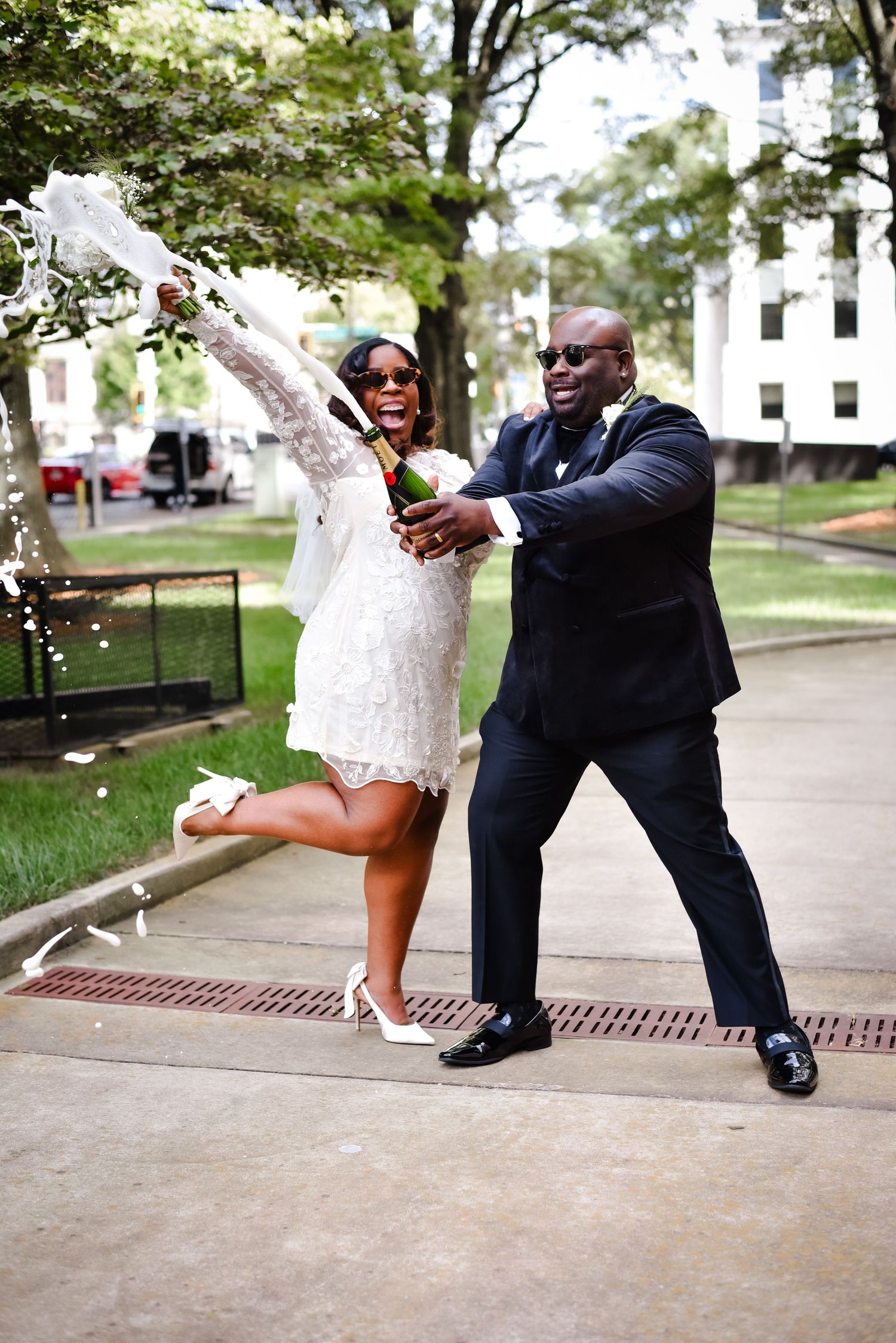 A couple in formal attire poses playfully near a bench in a park setting during a photo session.