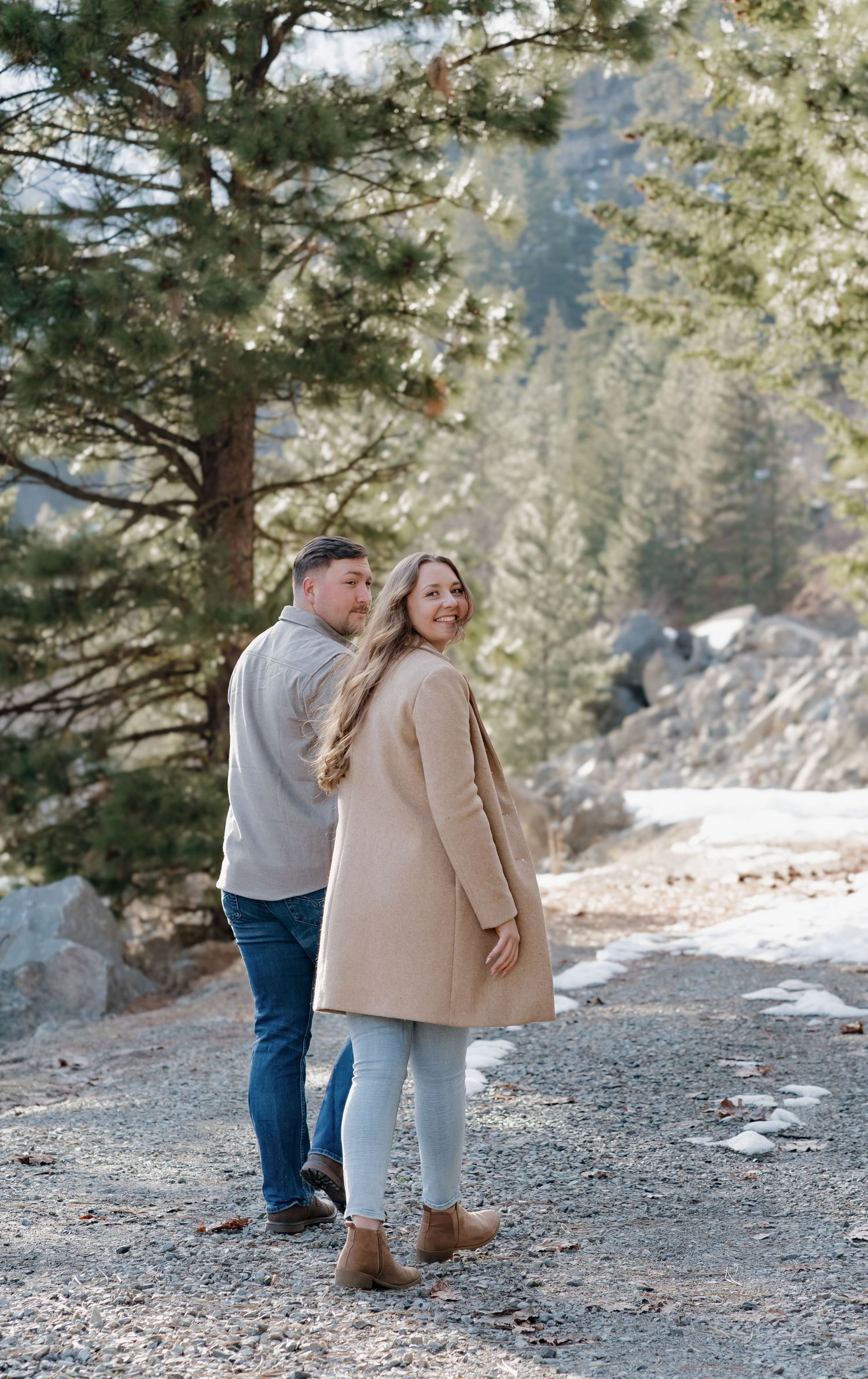 A couple walks together on a snowy mountain trail surrounded by evergreen trees on a winter day.
