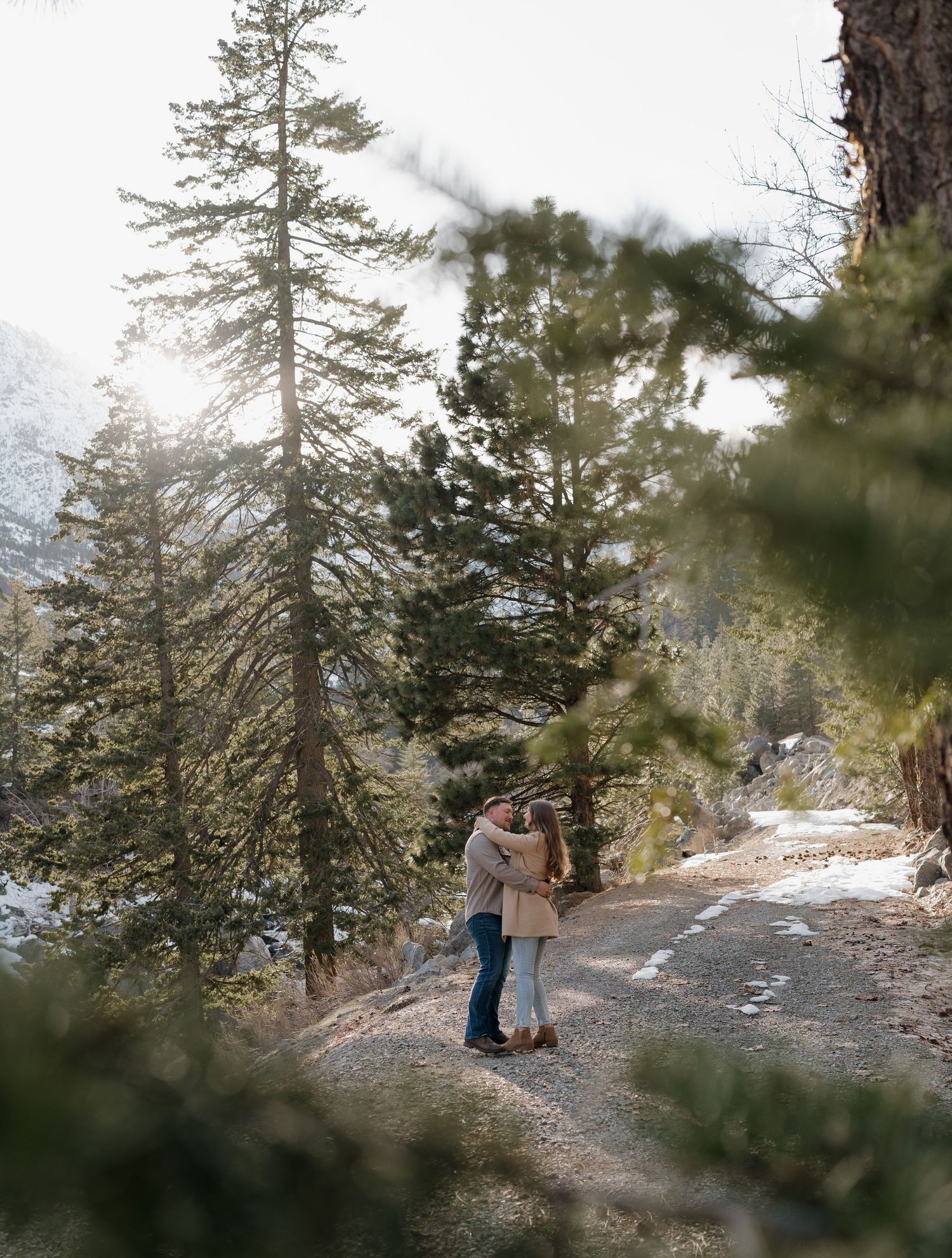A romantic moment captured between two people on a mountain path with pine trees and patches of snow.