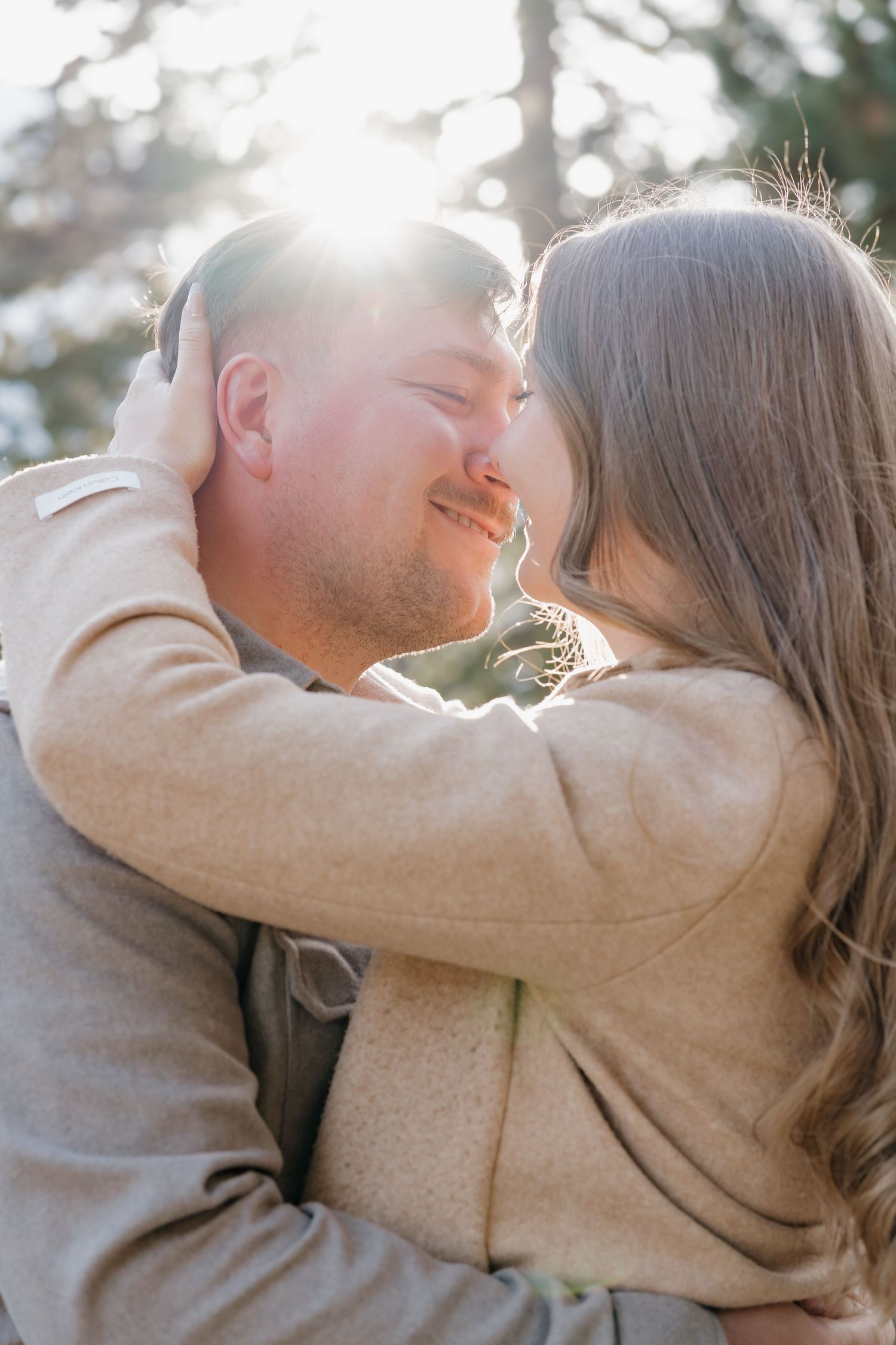 A tender embrace shared between a couple in warm winter clothing against a sunlit forest backdrop.