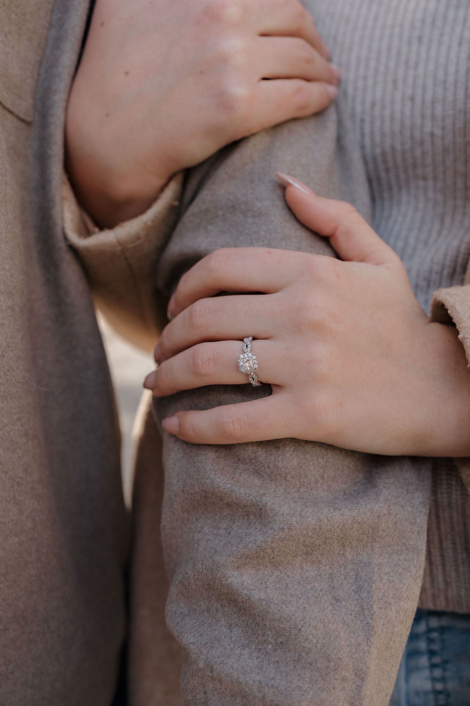 Close-up view of hands wearing a sparkling diamond engagement ring against a gray sweater background.