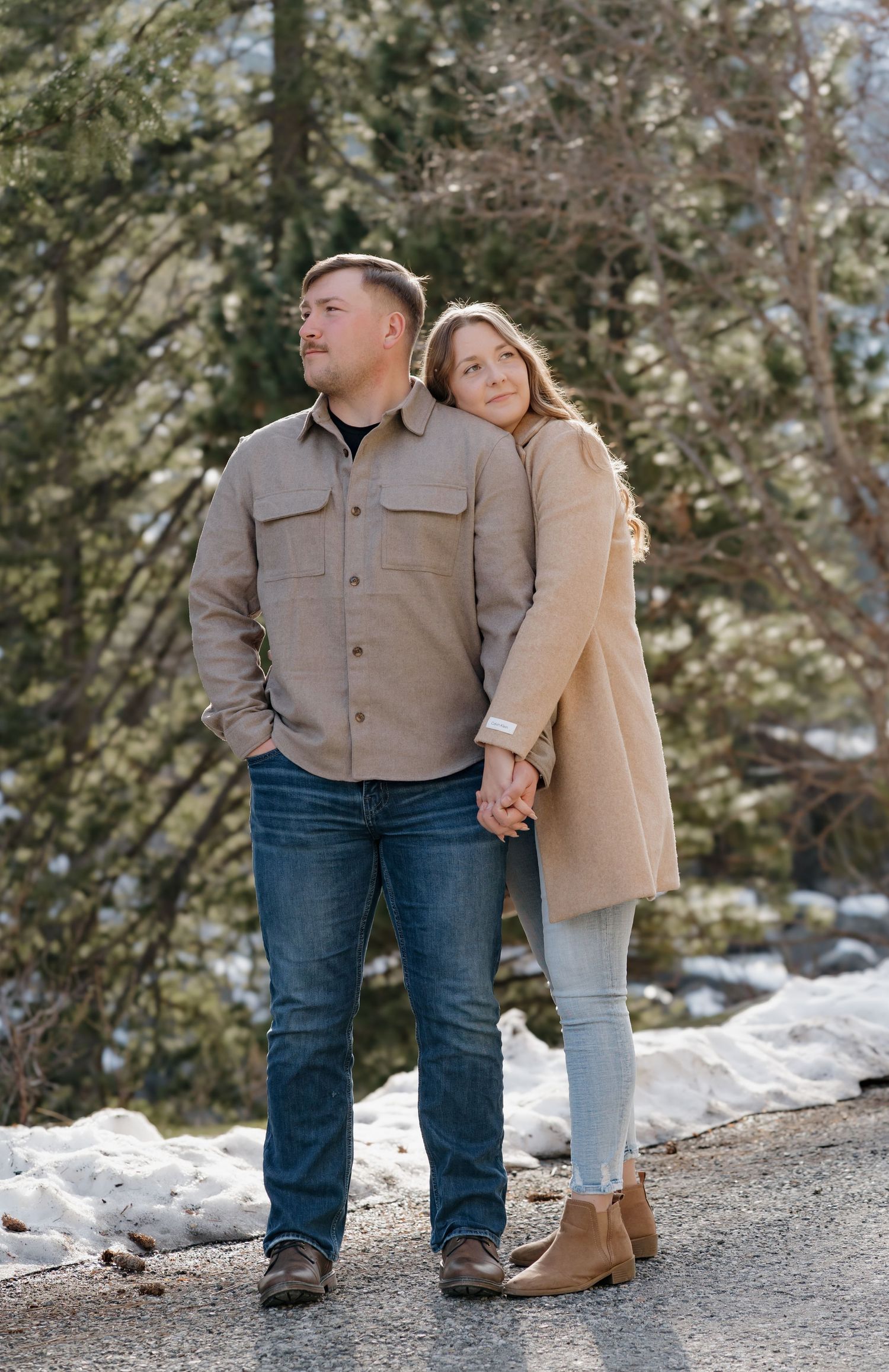A couple stands close together in a snowy winter setting wearing casual outerwear and jeans.