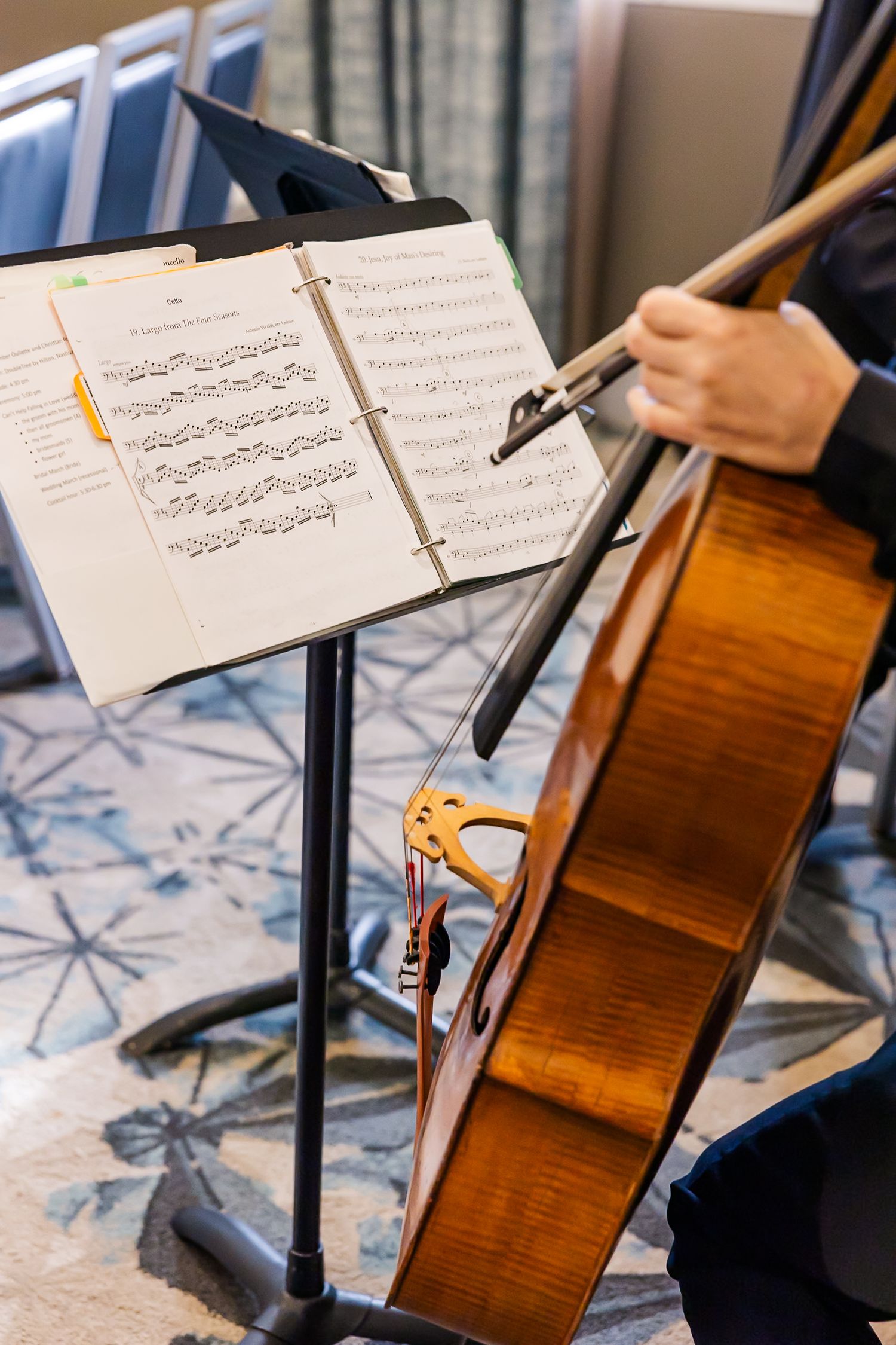 Cellist performs with sheet music on stand while playing wooden instrument in orchestra rehearsal setting.