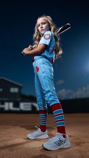 Baseball player wearing light blue uniform with red accents and white sneakers stands on dirt field at dusk.