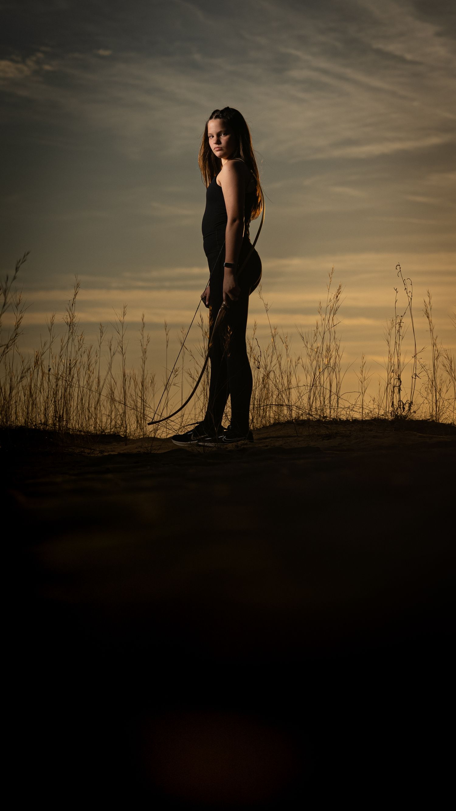 A silhouette stands in tall grass against a dramatic sunset sky wearing a dark outfit and long flowing hair.