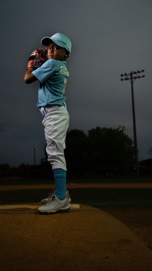 A young baseball player in teal uniform stands in pitching stance on a dirt mound at dusk with utility poles visible.