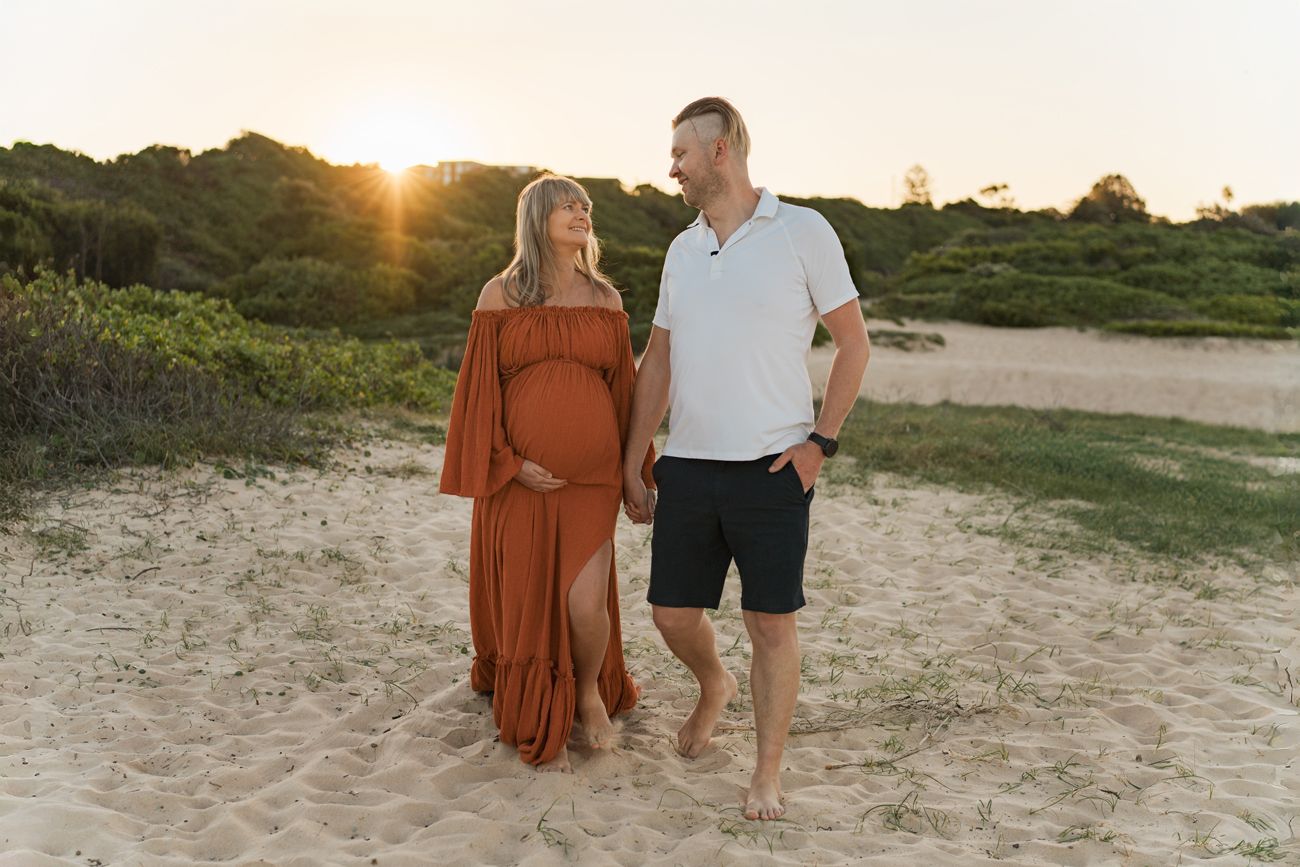 A couple walks along a sandy beach path during golden hour sunset as sunlight filters through trees maternity photoshoot in Sydney Maroubra