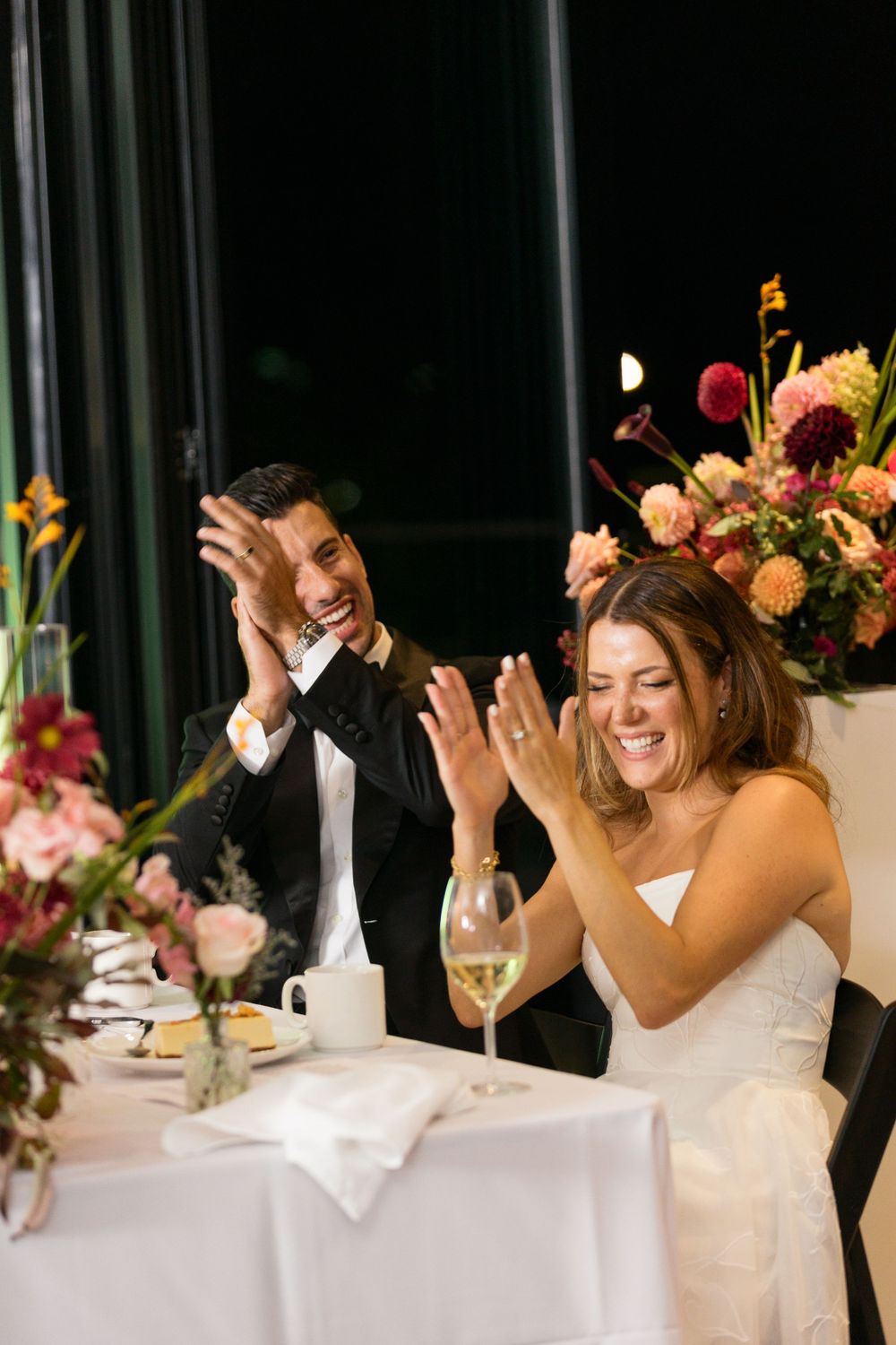 Wedding celebration at an elegant reception with joyful couple seated at table decorated with floral arrangements.
