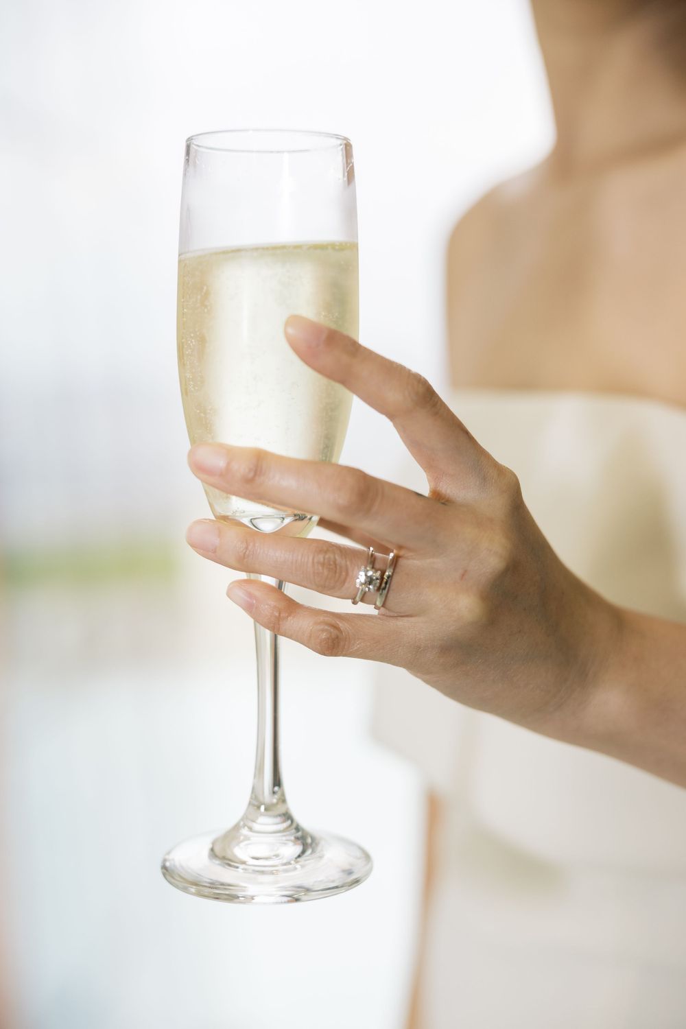 A hand wearing an engagement ring holds a champagne flute filled with sparkling wine against a light background.