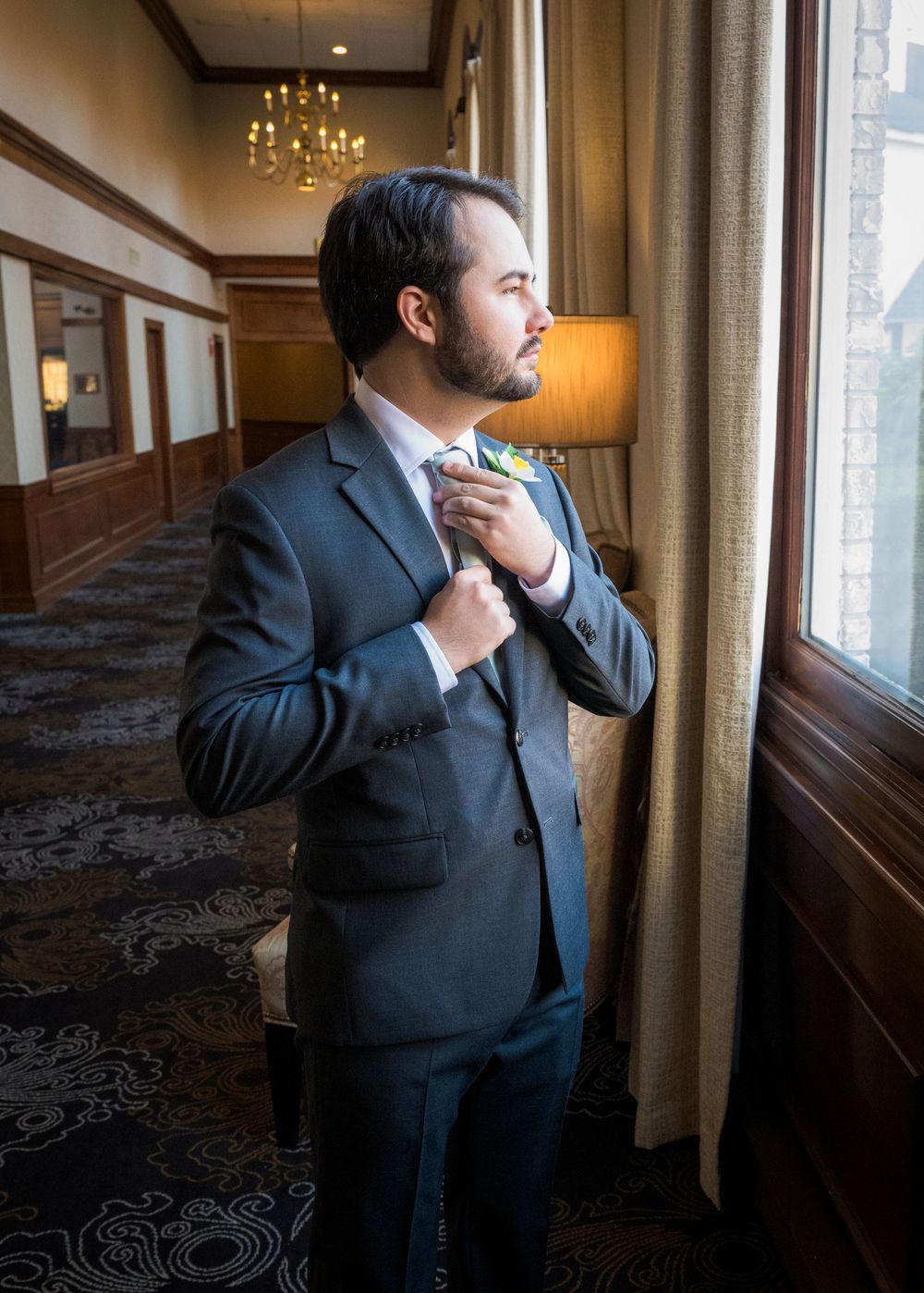 Groom in dark suit adjusts tie while looking out window in elegant hotel room.
