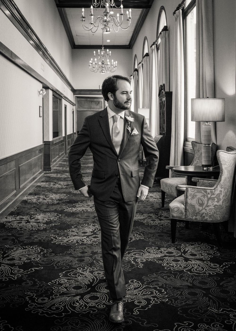 Black and white series of a groom walking through an elegant hallway with ornate carpet and chandeliers.