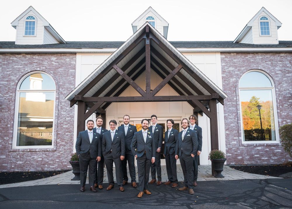Wedding party members pose together in formal attire outside a stone church building with white dormers.