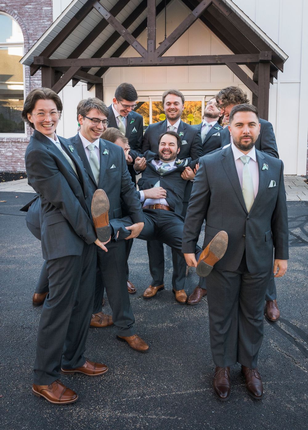 Groomsmen in dark suits posing playfully outside a brick building.