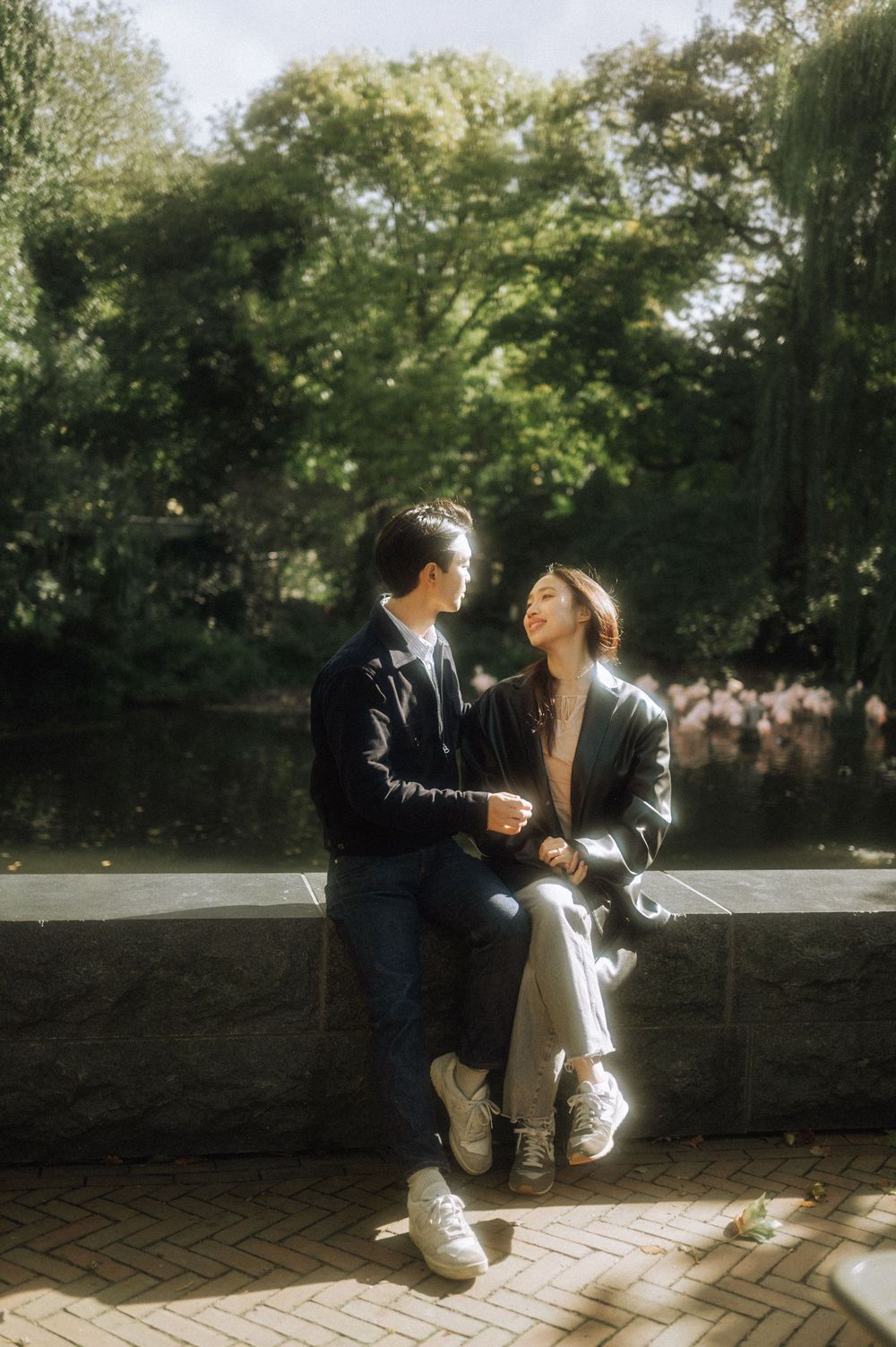 Two people share a tender moment on a stone wall path surrounded by sunlit trees.