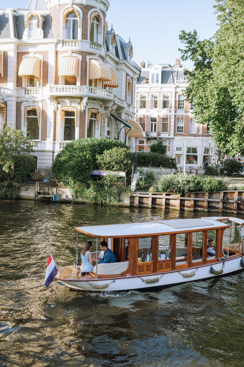 Wooden boat cruising along Amsterdam canal with historic buildings and trees in background.