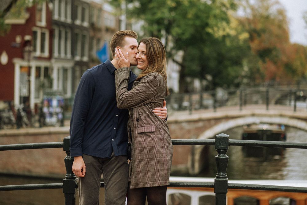 Couple embracing on autumn day along canal with historic buildings in background.