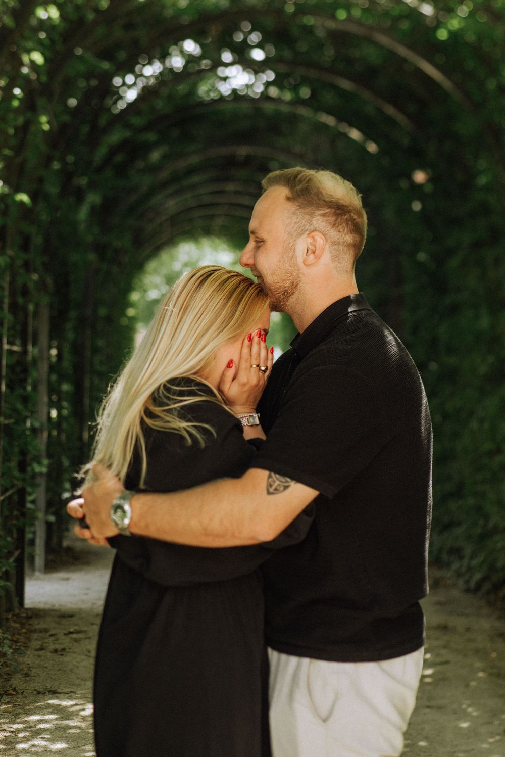 A couple embraces under a leafy green archway in a garden setting.
