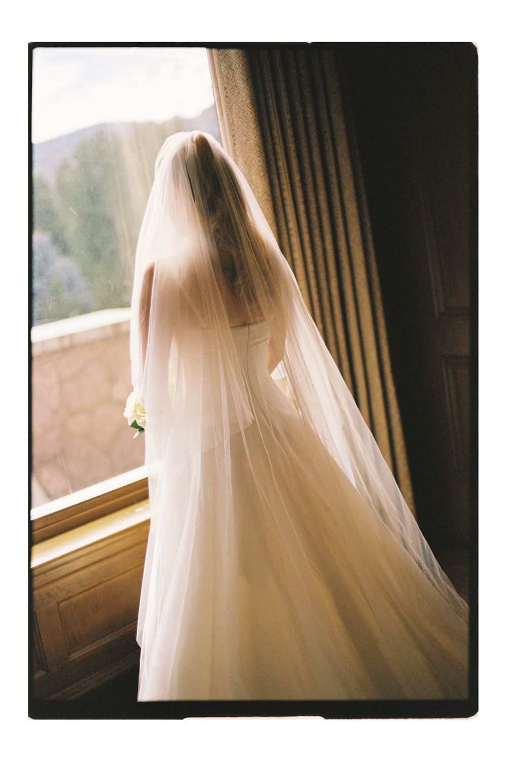 Bride in white wedding gown and long veil stands by sunlit window holding bouquet, viewed from behind.