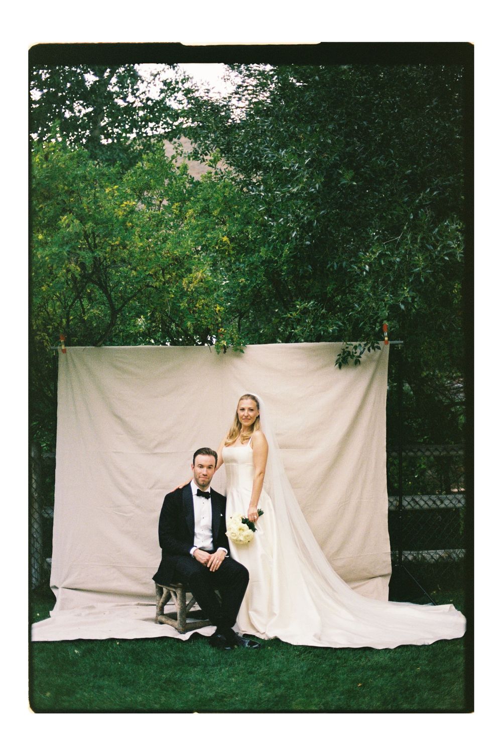 Bride in white gown stands behind groom in black tuxedo seated on stool before cream backdrop outdoors.