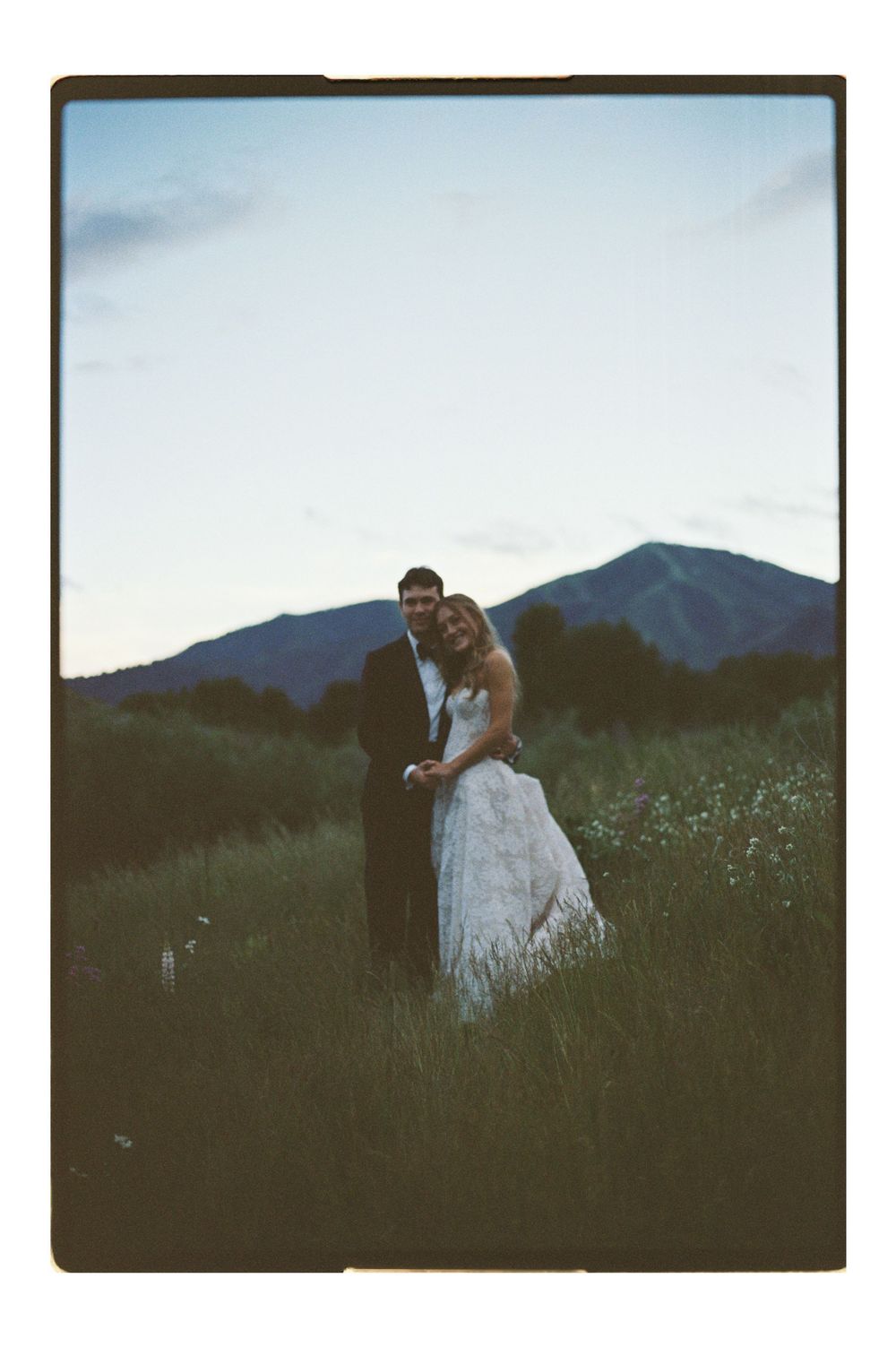Bride and groom embracing in a mountain meadow at dusk, wearing wedding attire in a romantic outdoor ceremony photo.